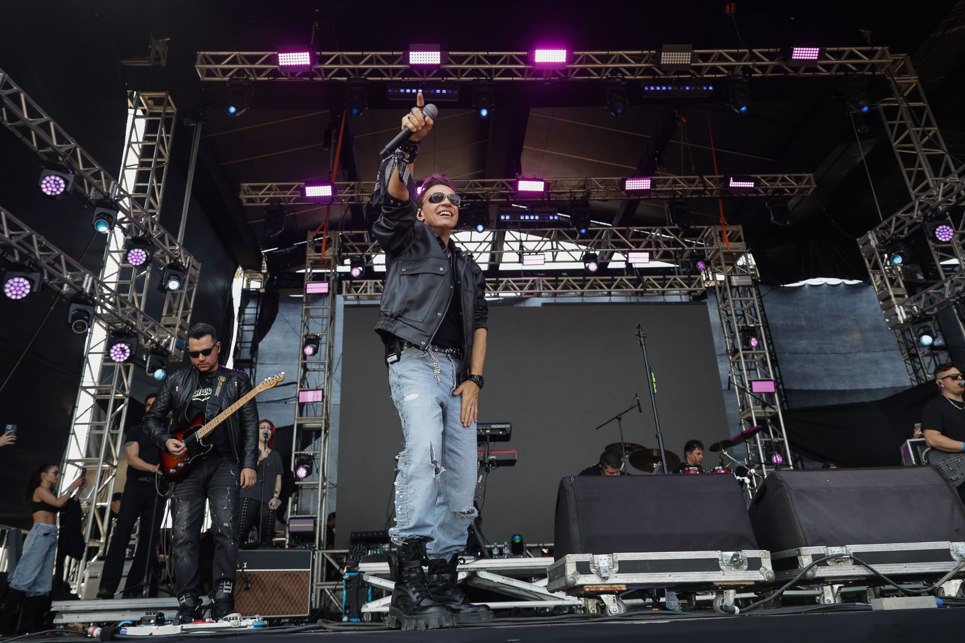 Um cantor de jaqueta de couro e óculos escuros se apresenta em um palco de shows com um guitarrista.