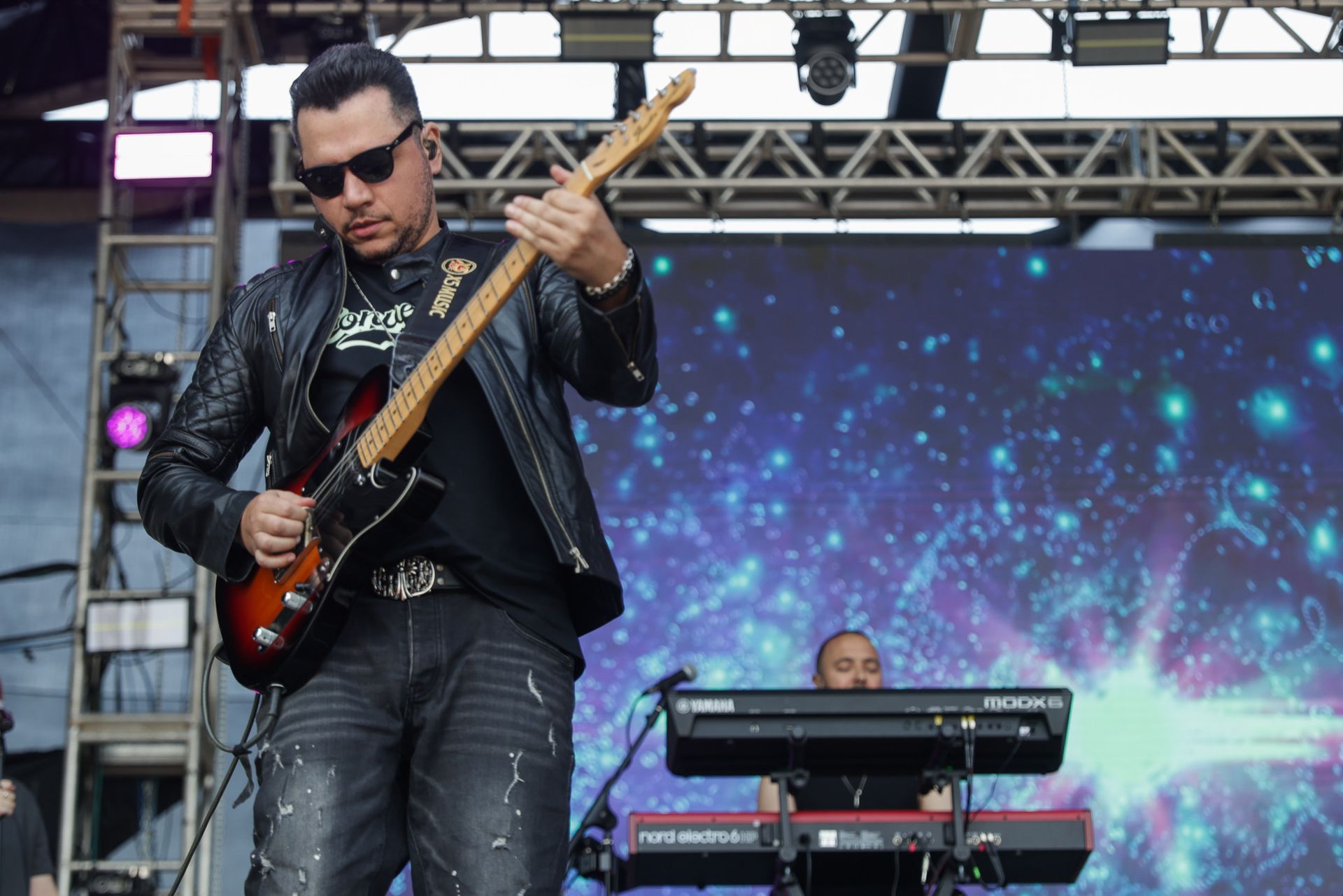 Um guitarrista de jaqueta de couro se apresenta no palco diante de um tecladista e um vibrante cenário digital.