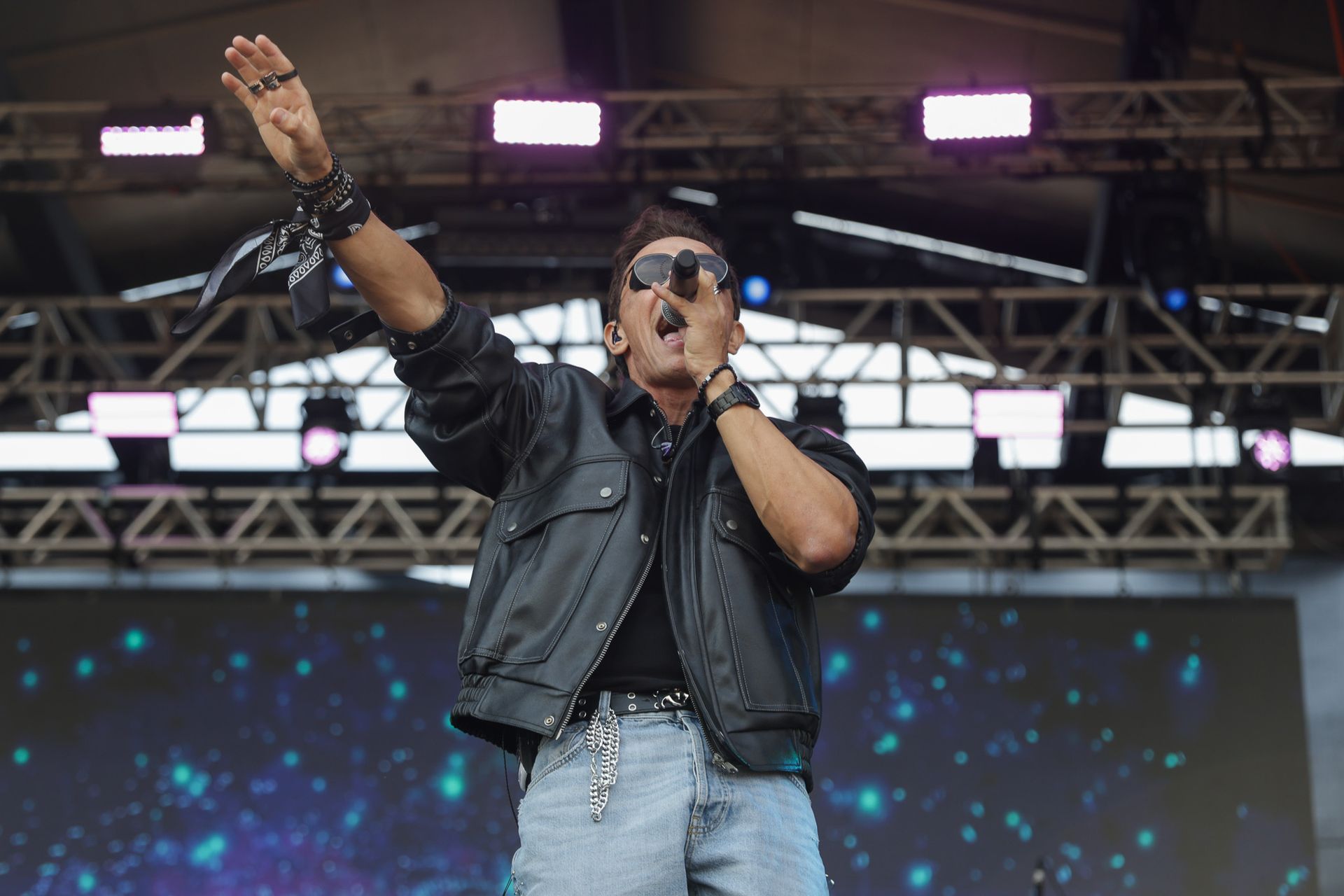 Um artista vestindo jaqueta de couro preta e óculos escuros em um palco com um fundo digital de luz azul.