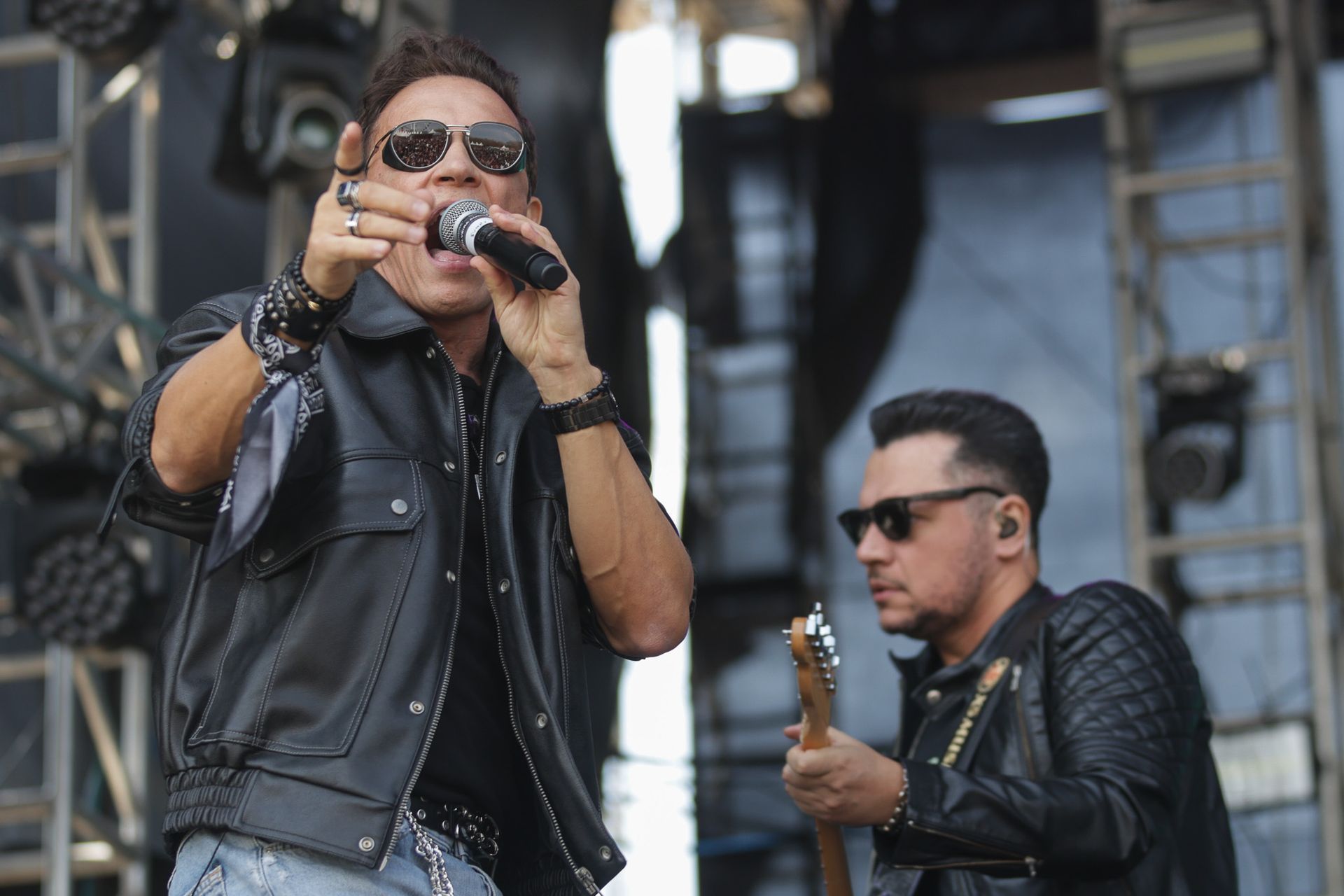 O vocalista aponta enquanto canta em um microfone, ao lado de um guitarrista no palco, com uma estrutura metálica ao fundo.