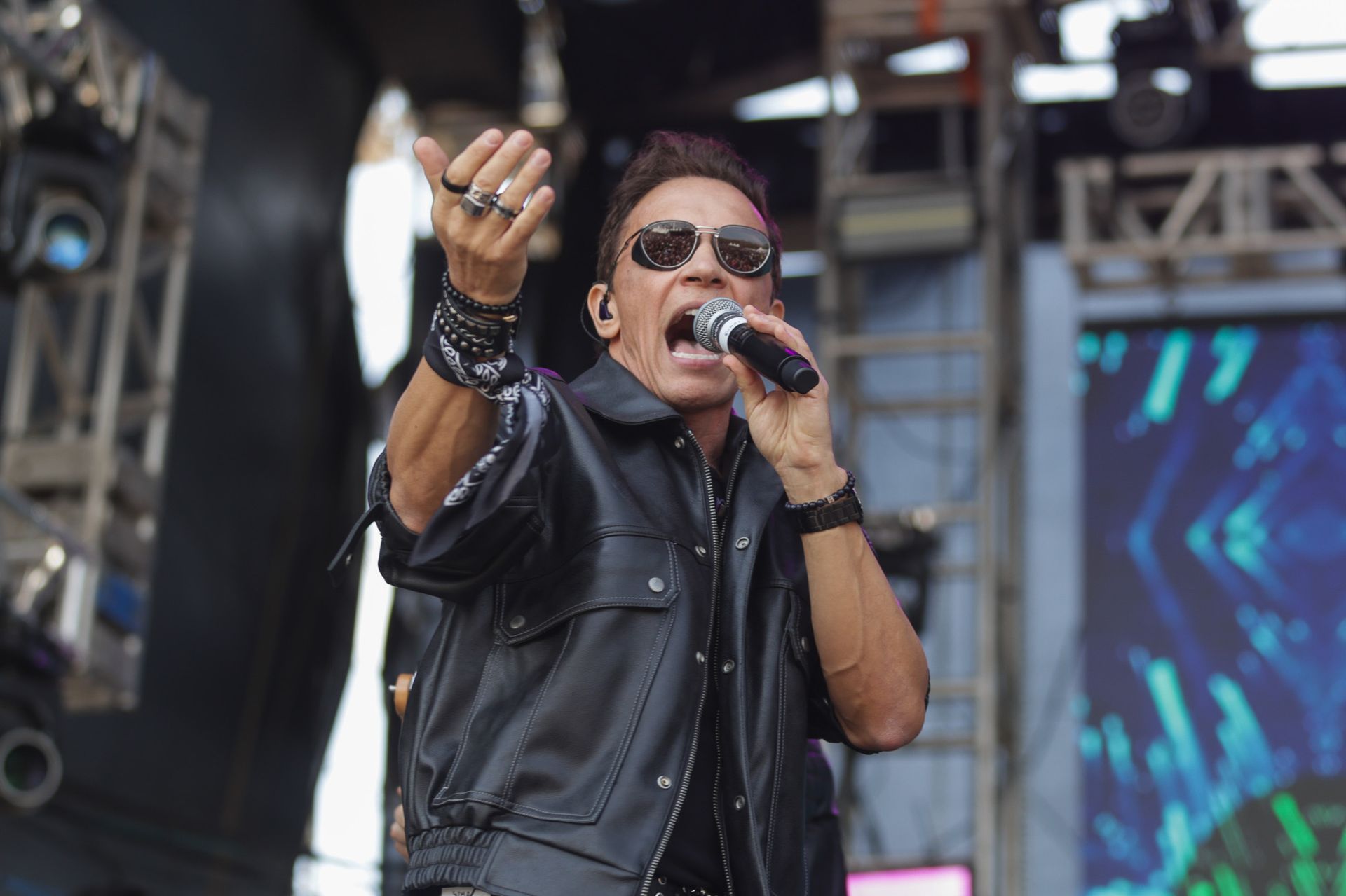 Um artista vestindo jaqueta de couro e óculos escuros canta em um microfone enquanto gesticula no palco de um show.