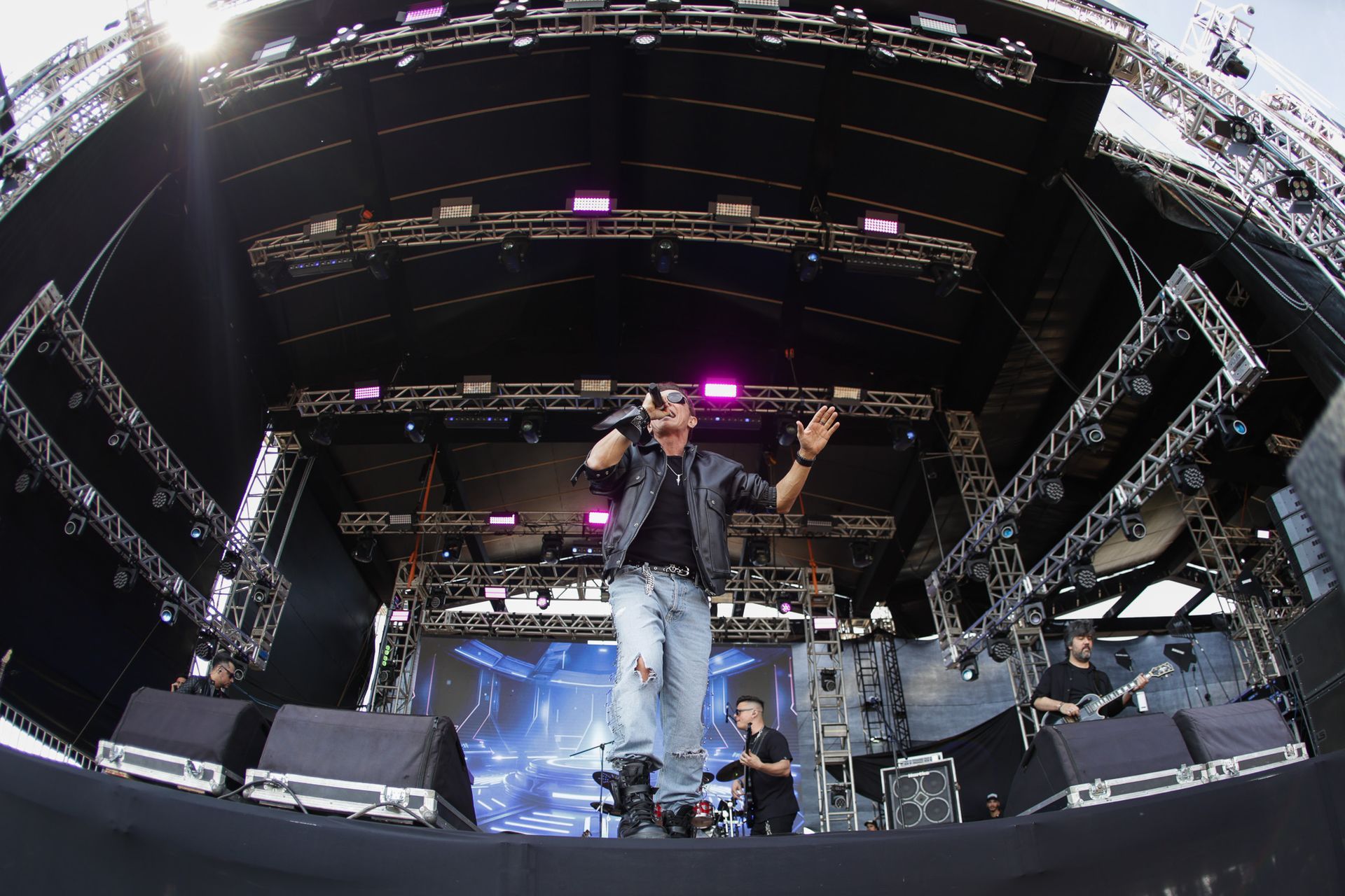 Um artista se apresenta em um grande palco ao ar livre sob luzes brilhantes, cantando em um microfone com o braço estendido.