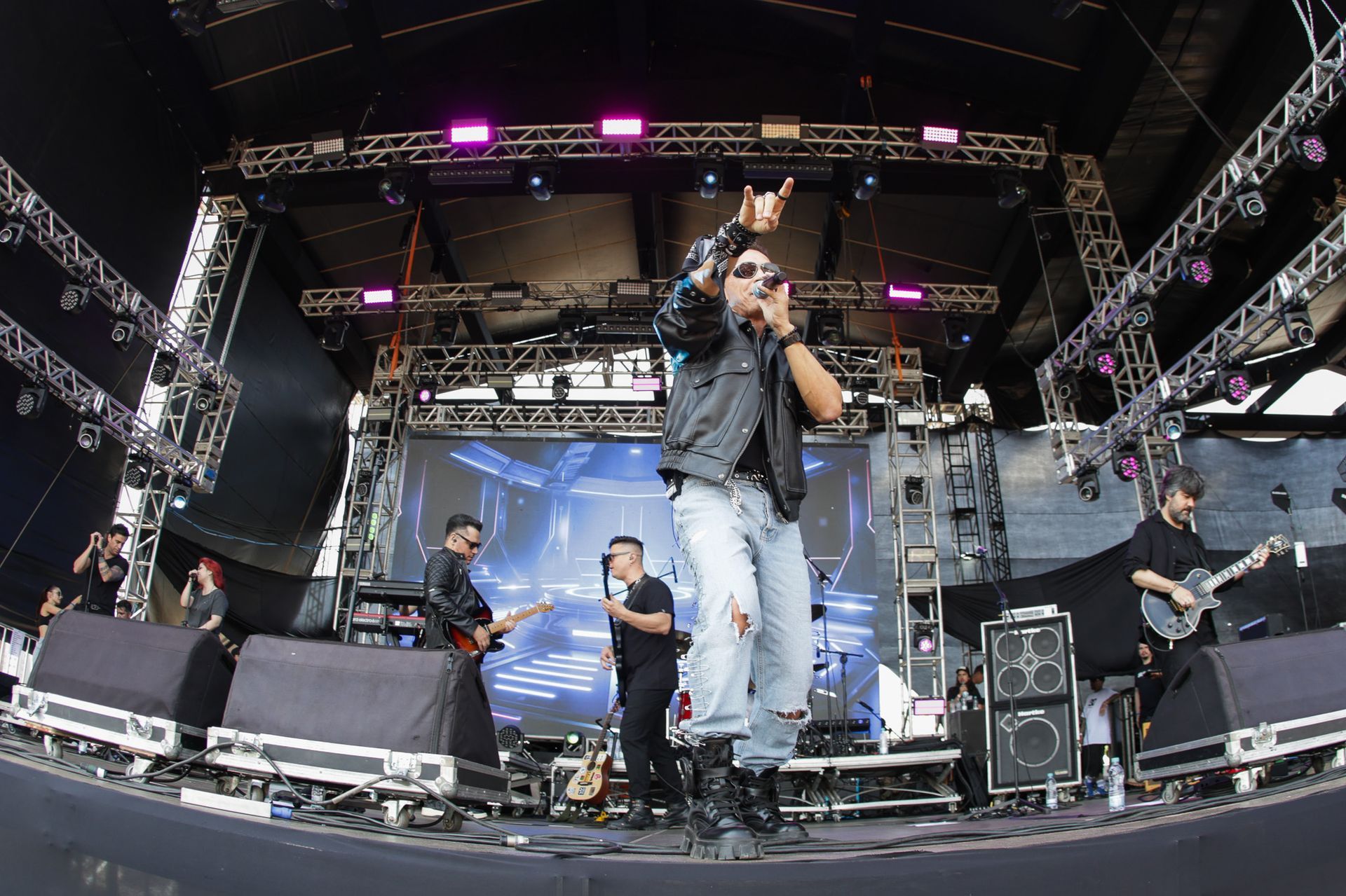 Uma banda se apresentando em um palco ao ar livre, com um vocalista principal e músicos acompanhantes.