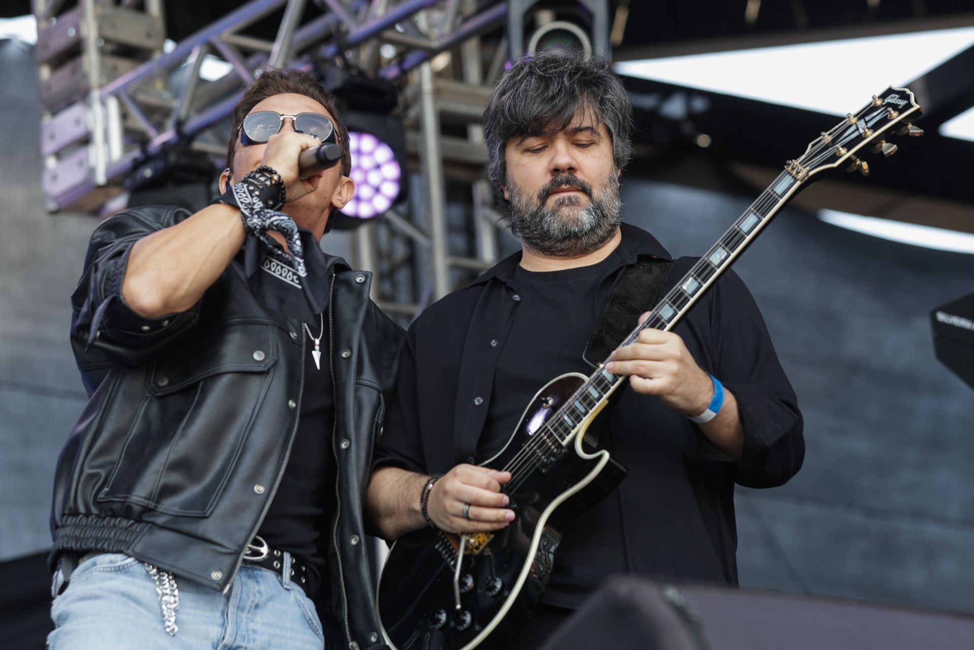 Um cantor de óculos escuros e jaqueta de couro se apresenta ao lado de um guitarrista 