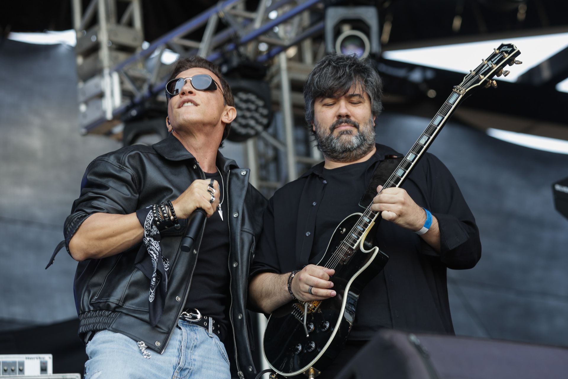 Um cantor de óculos escuros e jaqueta de couro se apresenta no palco acompanhada por um guitarrista.