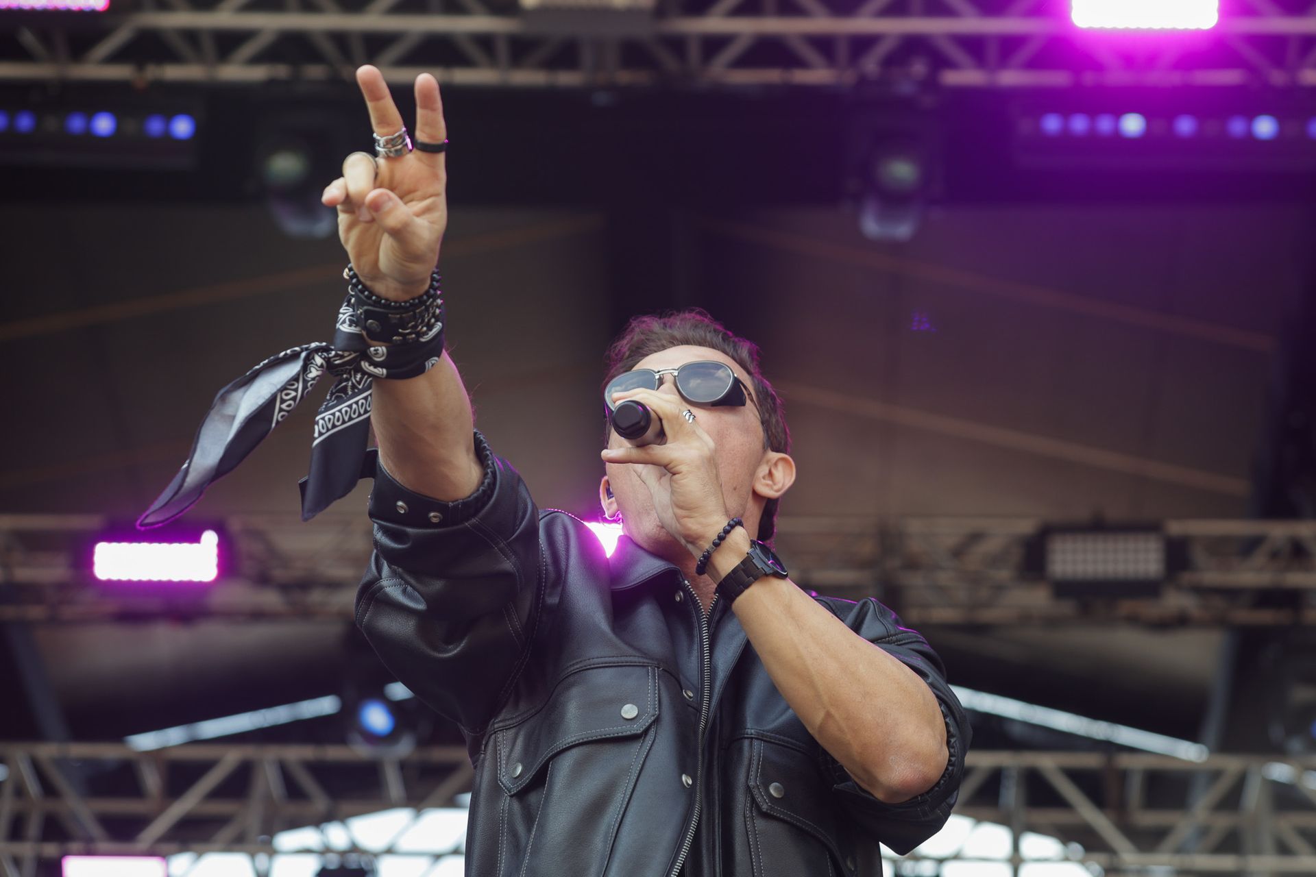 Um artista vestindo jaqueta de couro e óculos escuros canta em um microfone no palco, fazendo o sinal de paz com os dedos.