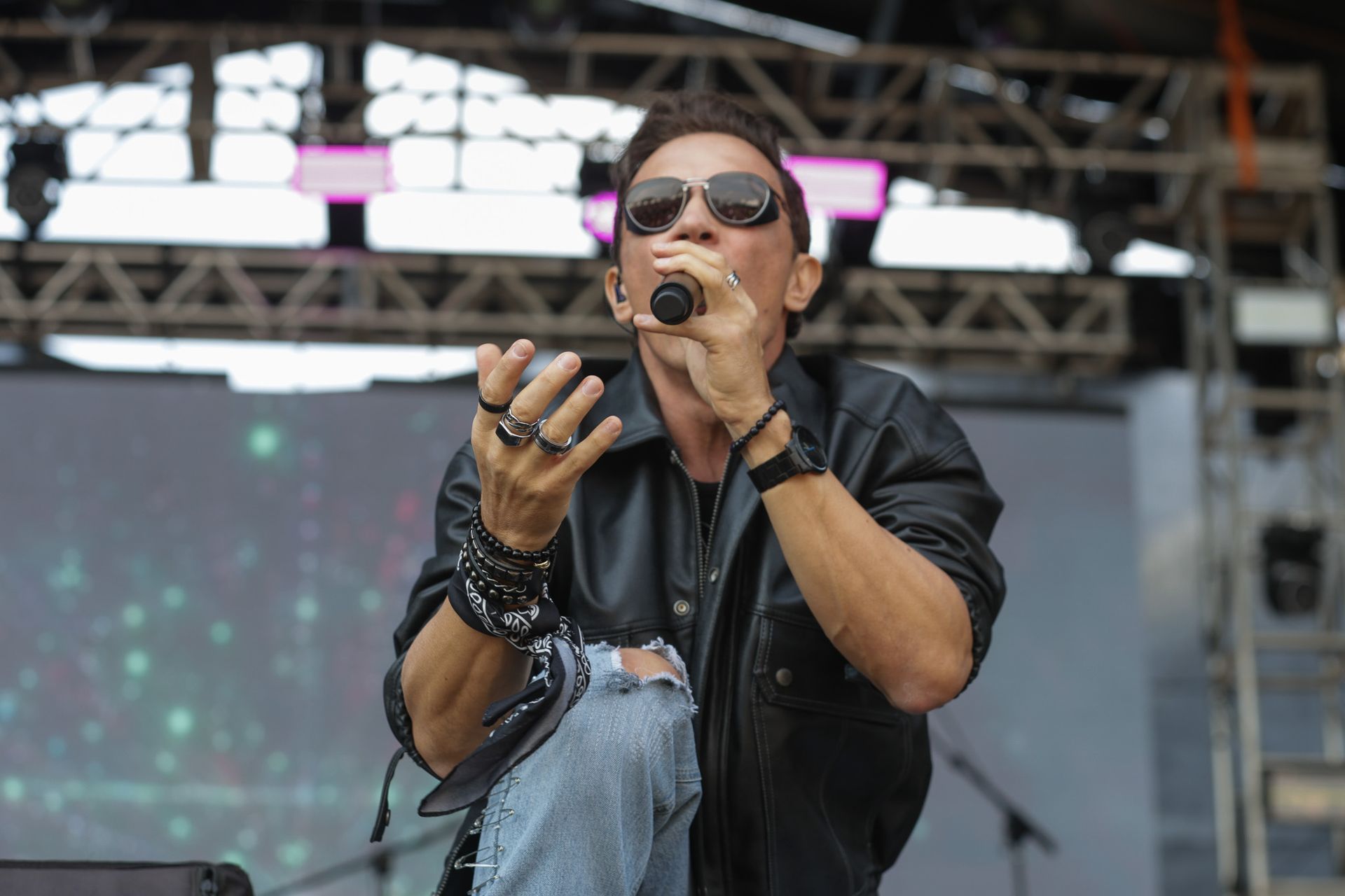 Um artista vestindo jaqueta de couro e óculos escuros canta em um microfone no palco, gesticulando com anéis nos dedos.