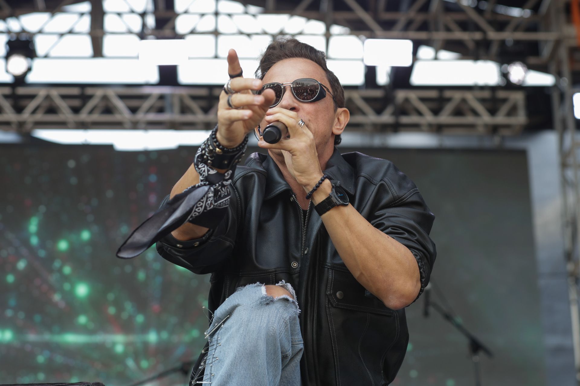 Um artista vestindo jaqueta de couro preta e óculos escuros canta enquanto está ajoelhado no palco de um show.