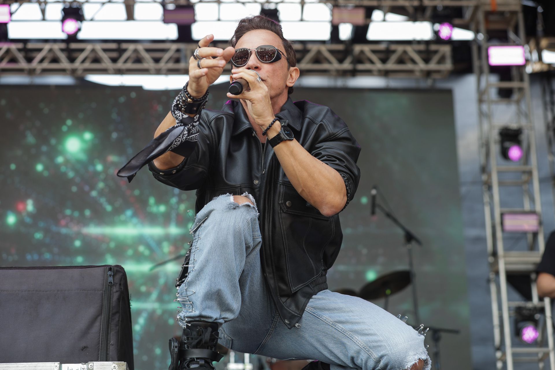 Um artista vestindo jaqueta de couro preta e calça jeans azul-clara rasgada, agachado enquanto canta no palco.