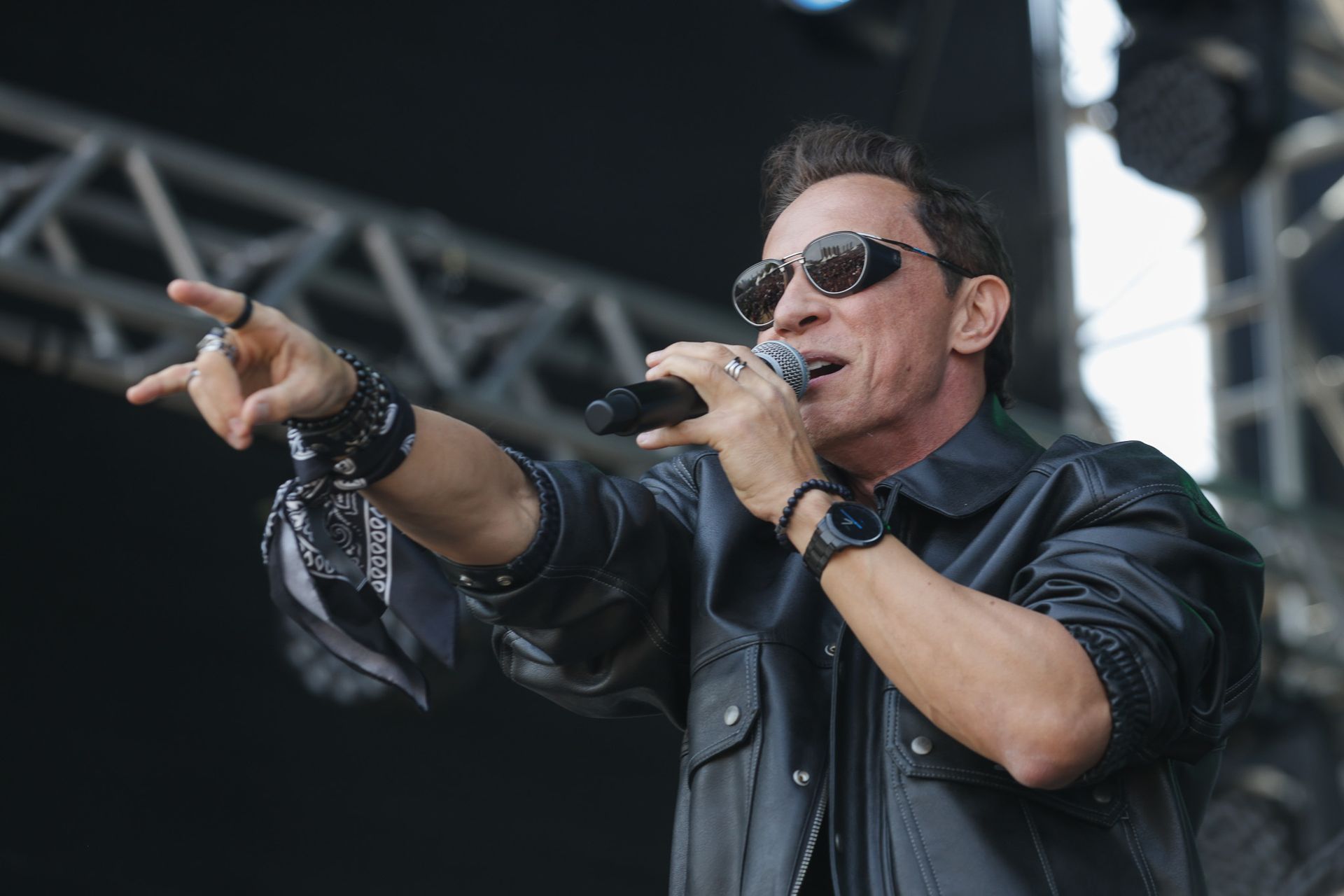 Um artista vestindo jaqueta de couro preta e óculos escuros canta em um microfone enquanto aponta para a plateia.