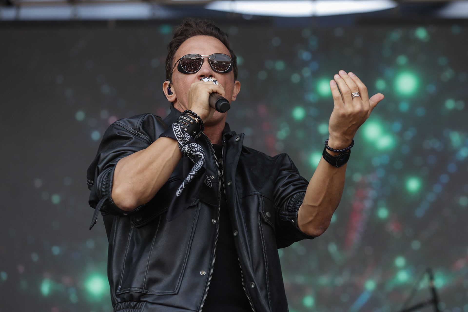 Um artista vestindo jaqueta de couro preta e óculos escuros canta em um microfone no palco contra um fundo verde desfocado.