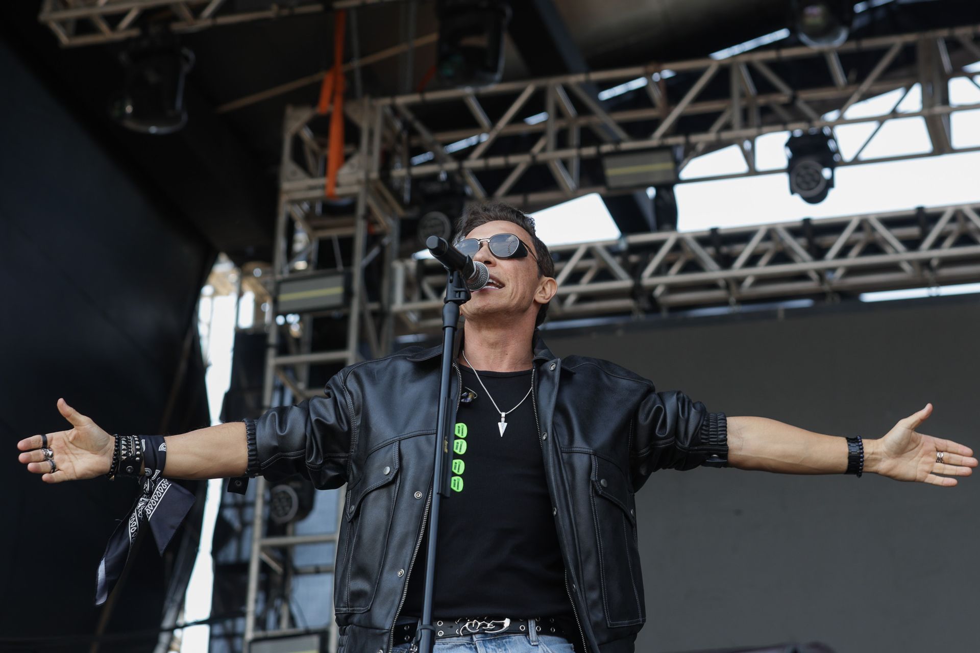 Um artista  vestindo jaqueta de couro e óculos escuros canta com os braços estendidos em um palco de shows.
