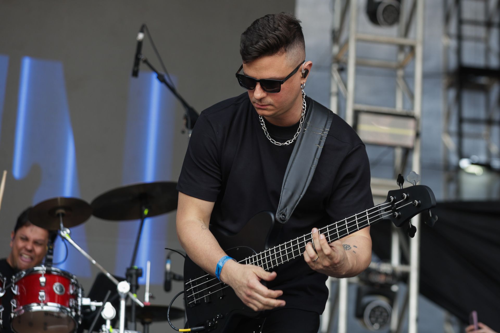 Um músico toca um baixo preto no palco, com um baterista visível ao fundo.