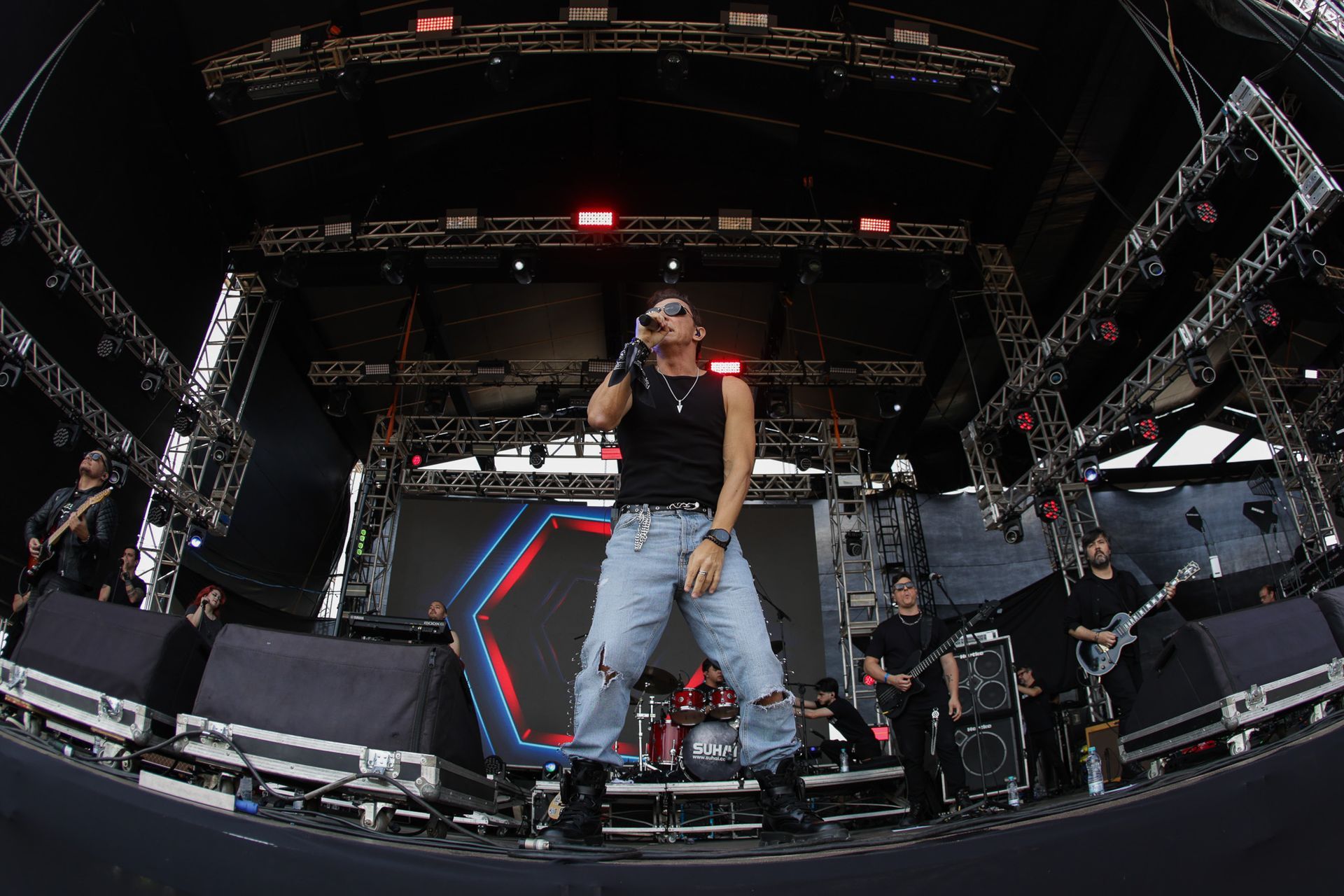Um artista vestindo uma regata preta e calça jeans azul-clara rasgada cantando no palco de um show com uma banda.