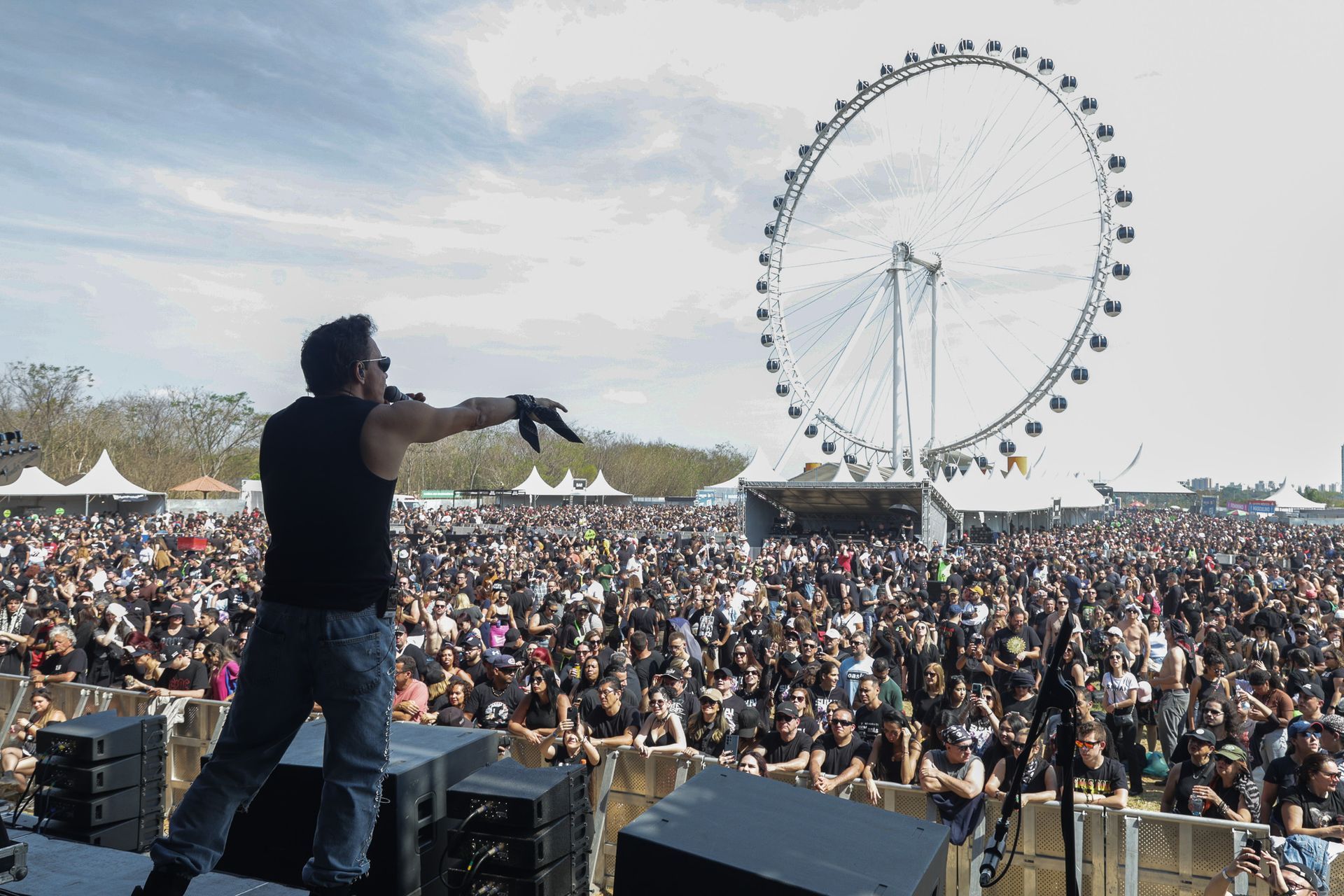 Um artista no palco, uma grande multidão que o aplaude em um festival ao ar livre, com uma roda-gigante ao fundo.