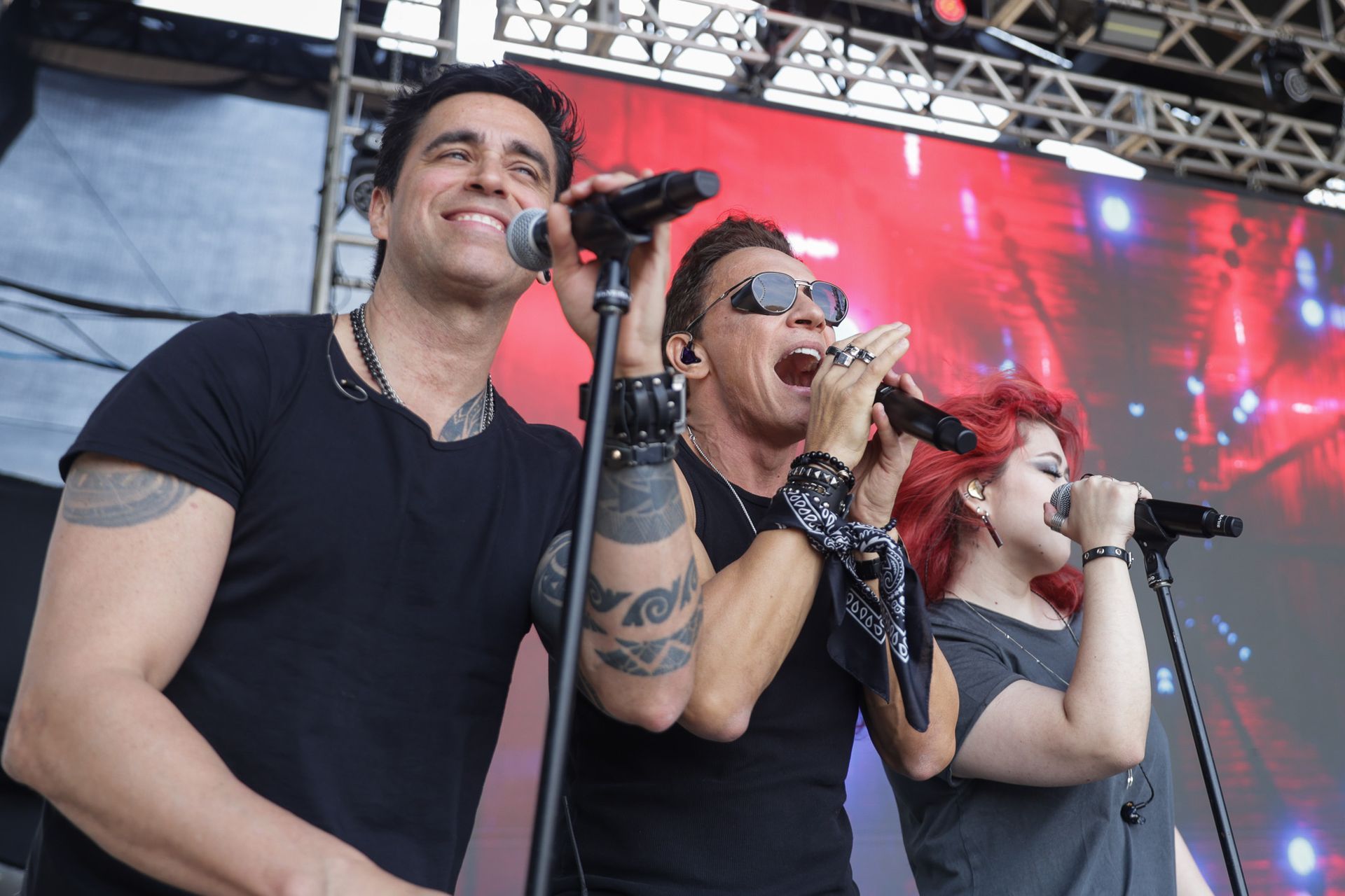Três cantores de camisa preta se apresentam juntos no palco, segurando microfones e cantando para a plateia.