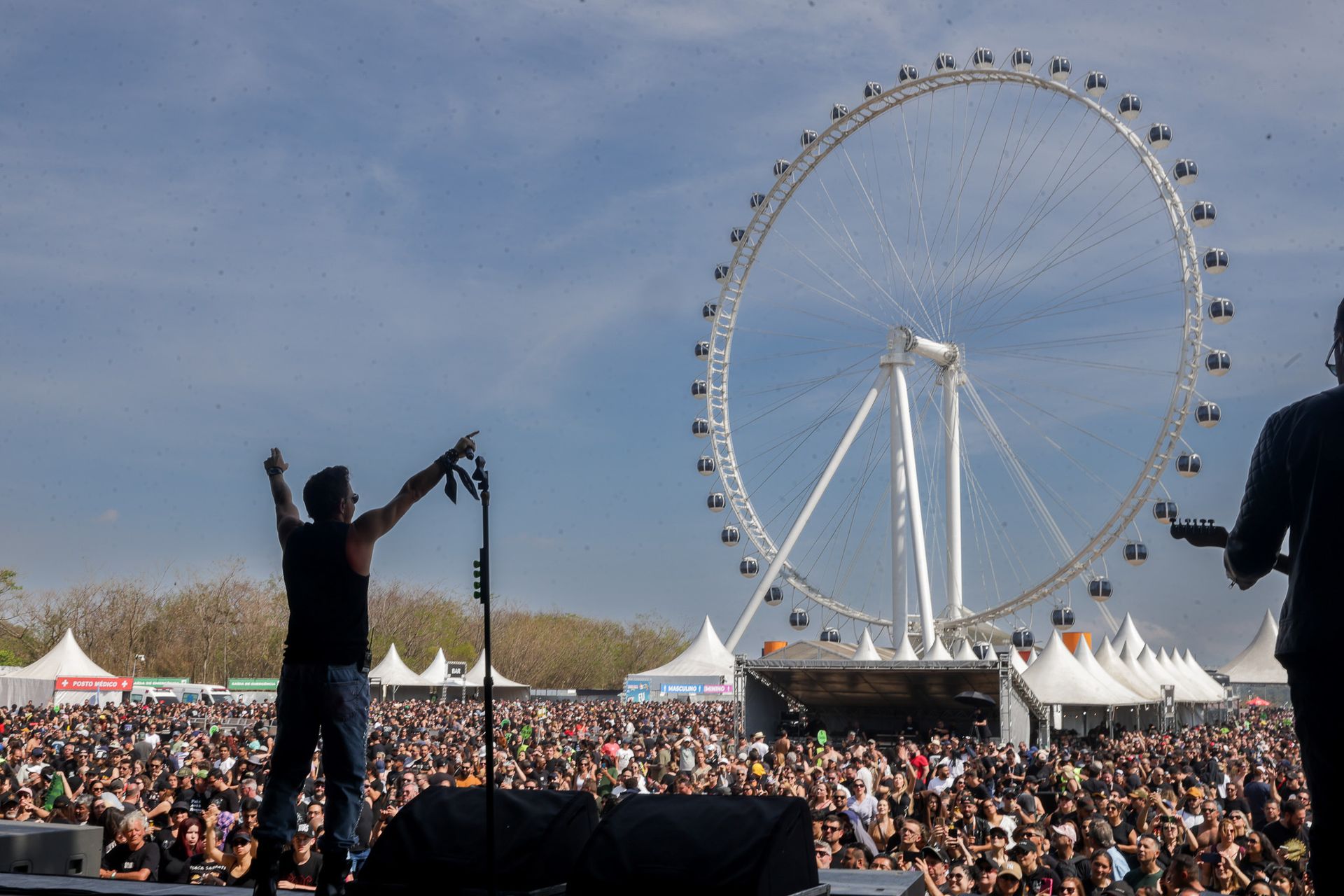 Um artista no palco com os braços erguidos, de frente para uma grande multidão e uma roda-gigante sob um céu azul claro.