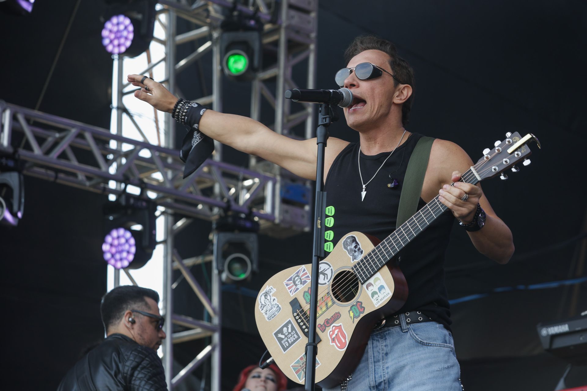 Um músico de regata preta e óculos escuros canta ao microfone enquanto toca violão em um palco de concerto.