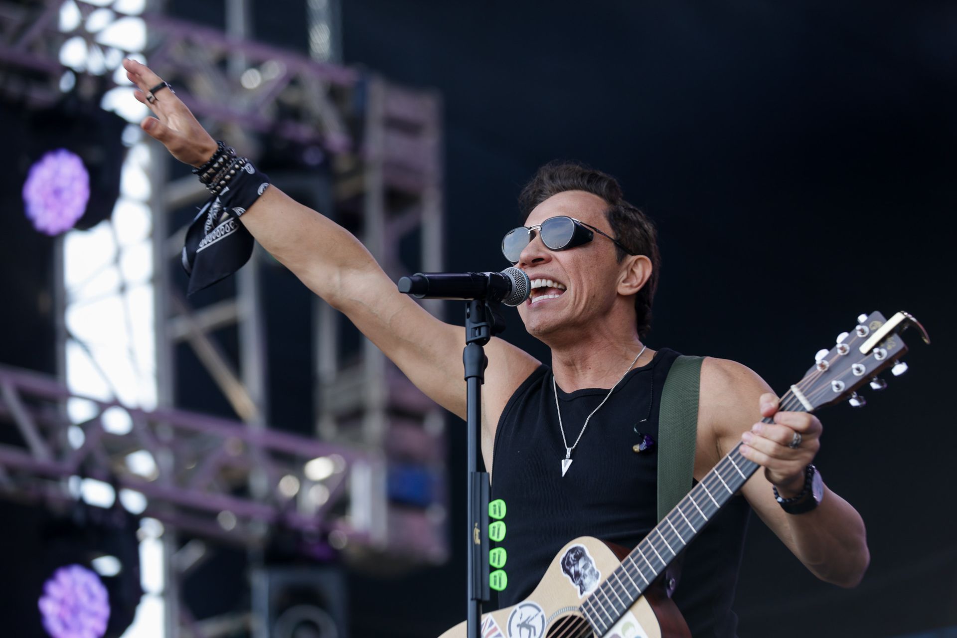 Um músico de óculos escuros e regata preta se apresenta no palco, com o braço levantado enquanto toca violão.