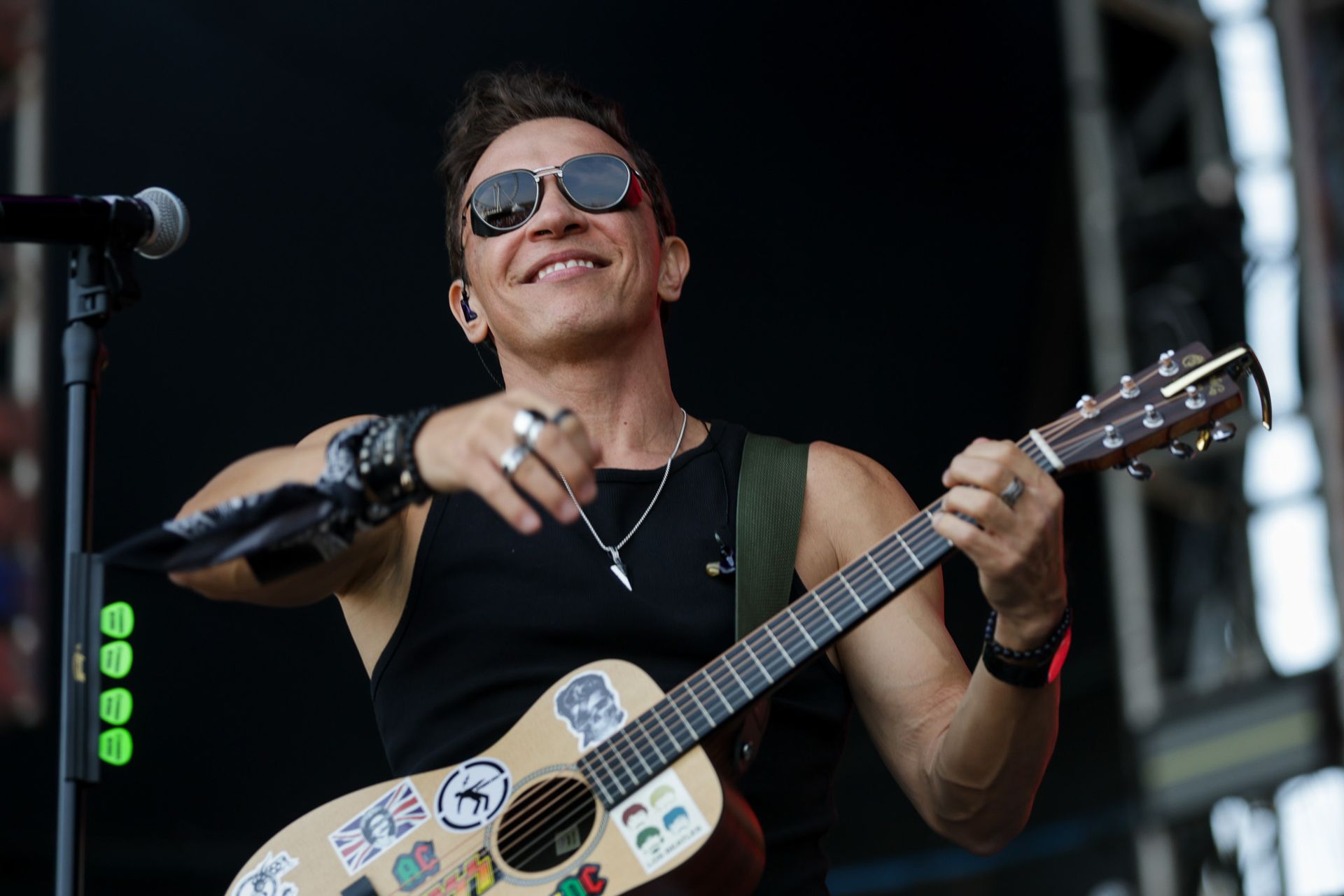 Um artista usando óculos escuros e uma regata toca um violão coberto de adesivos em um palco de shows.