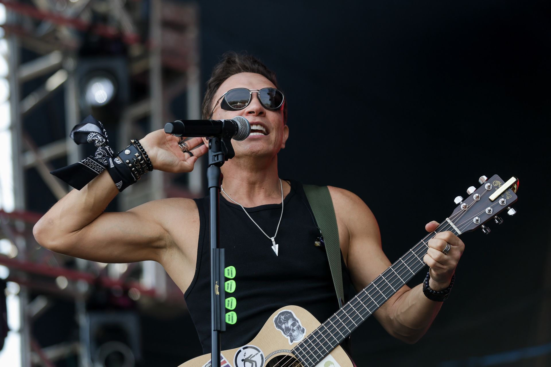 Um artista de óculos escuros cantando ao microfone enquanto toca violão em um palco com equipamentos de som para shows.