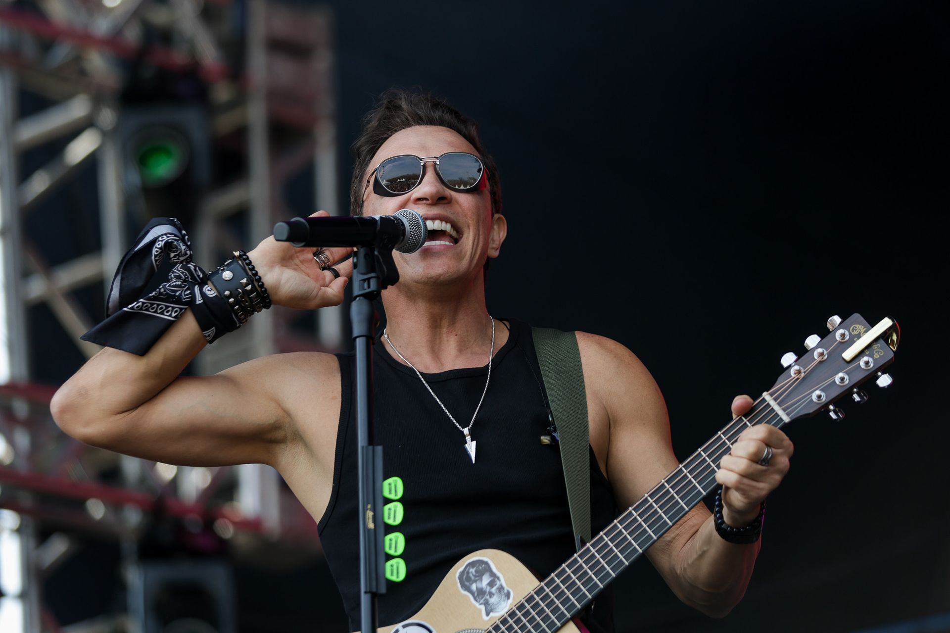 Um artista usando óculos escuros e uma regata preta canta em um microfone enquanto toca violão no palco.