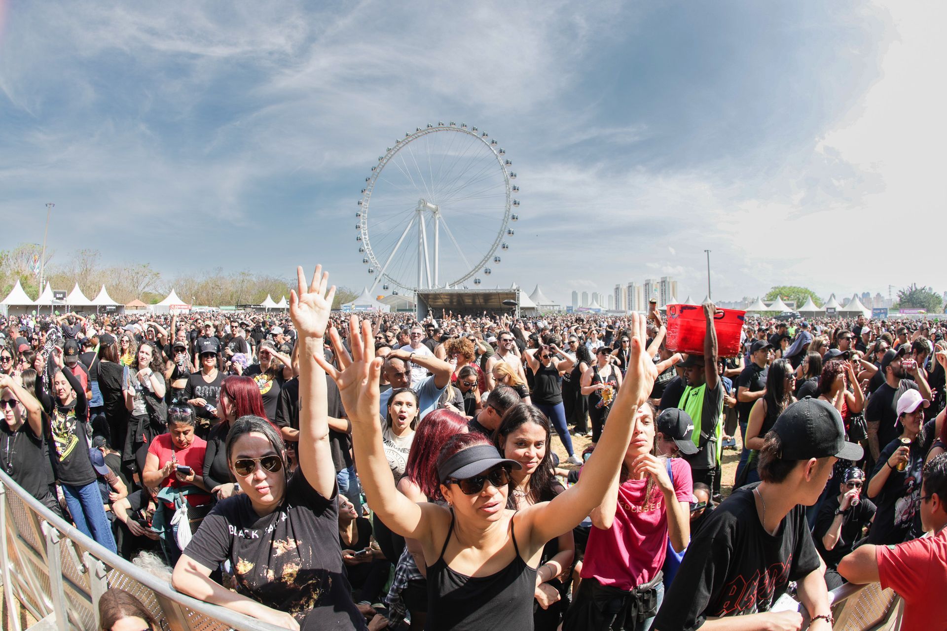 Uma multidão em um festival ao ar livre vibra com os braços erguidos, com uma grande roda-gigante visível ao longe.