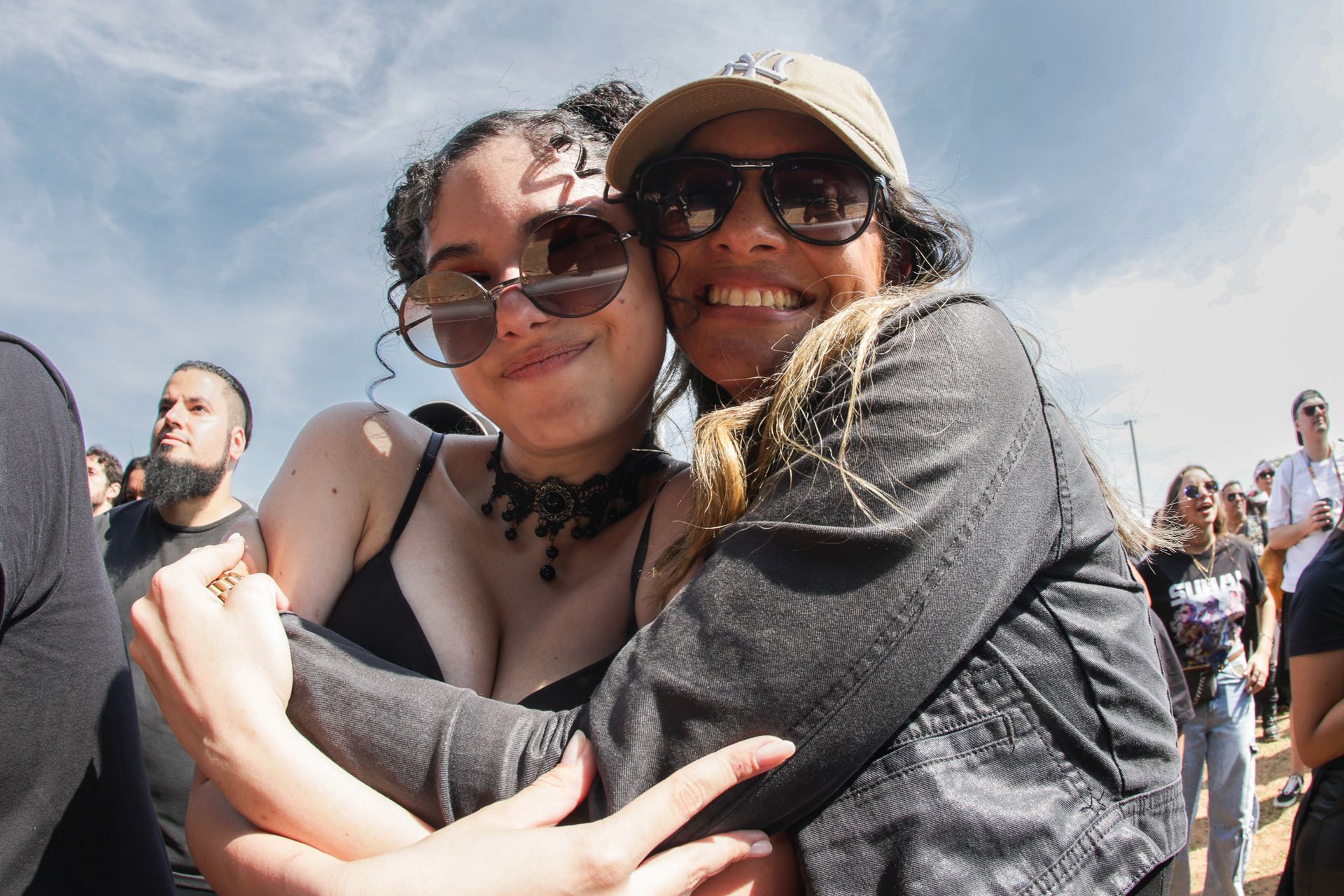 Duas pessoas se abraçam em um evento ao ar livre, usando óculos de sol e roupas casuais sob um céu claro e ensolarado.