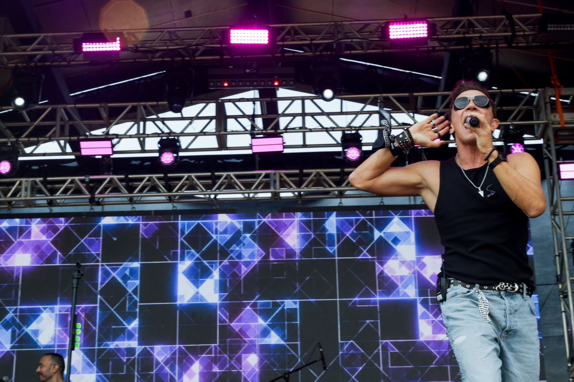 Um artista  vestindo uma regata preta e óculos escuros canta em um microfone em um palco com uma tela geométrica roxa.