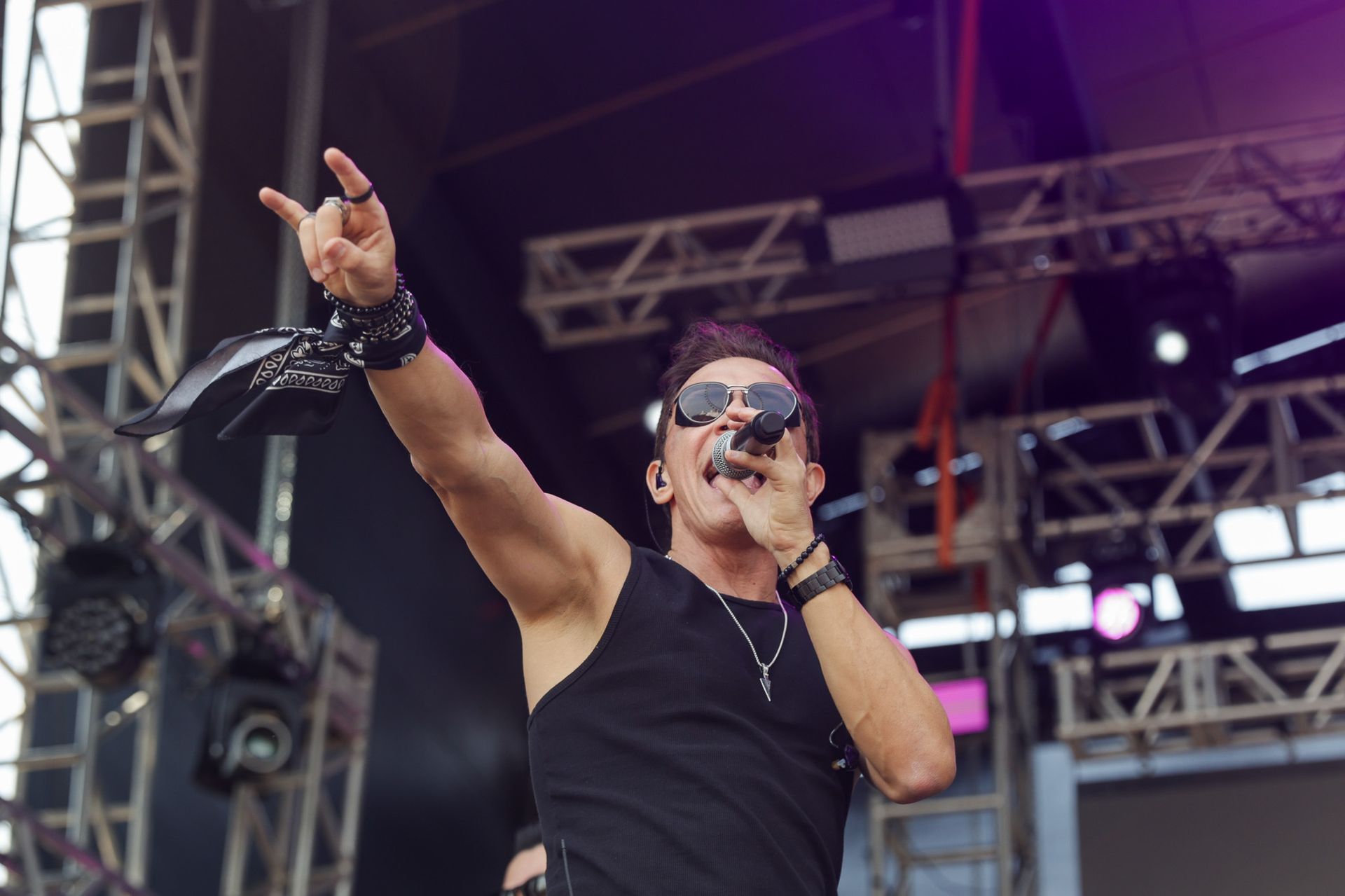Um artista vestindo uma regata preta e óculos escuros canta em um microfone no palco, fazendo um gesto de 