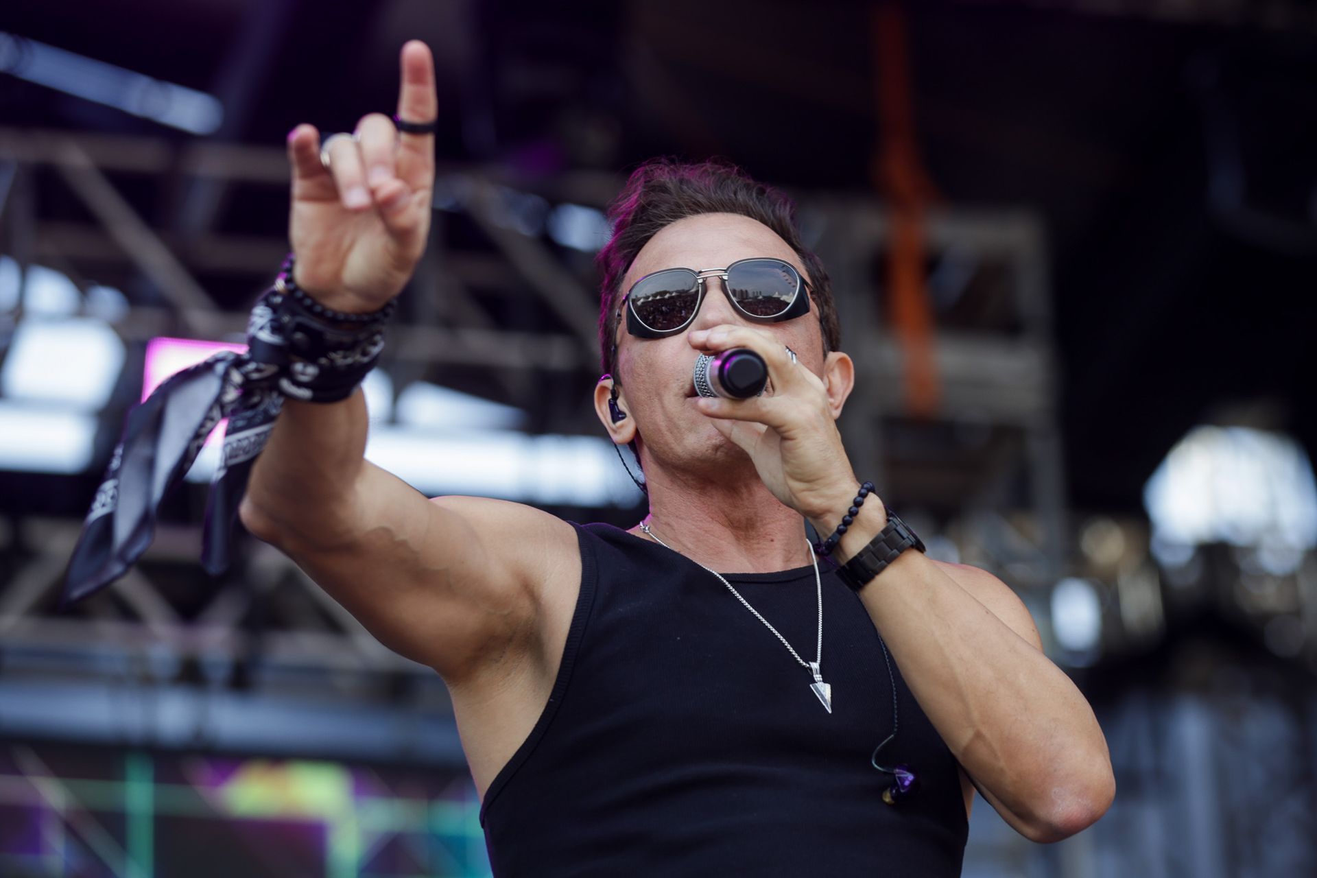 Um músico de óculos escuros e regata preta canta em um microfone enquanto gesticula no palco de um show.