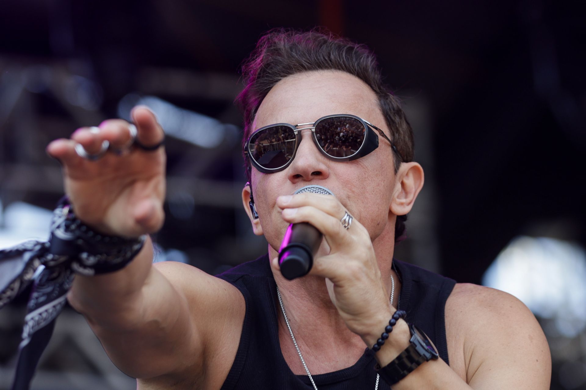 Um artista usando óculos escuros canta no palco, estendendo um braço para a frente com a mão envolta em uma bandana.