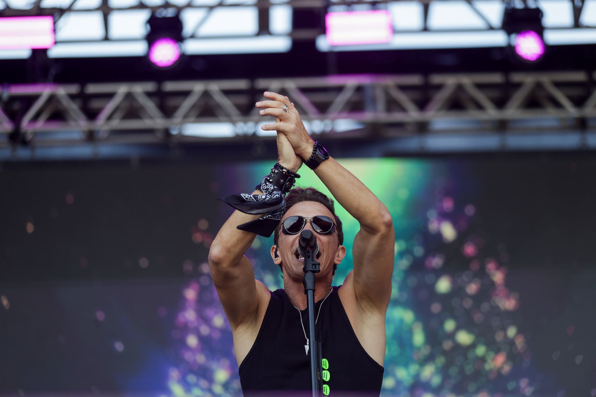 Um artista usando óculos escuros bate palmas enquanto está em um palco em frente a uma tela colorida e abstrata.