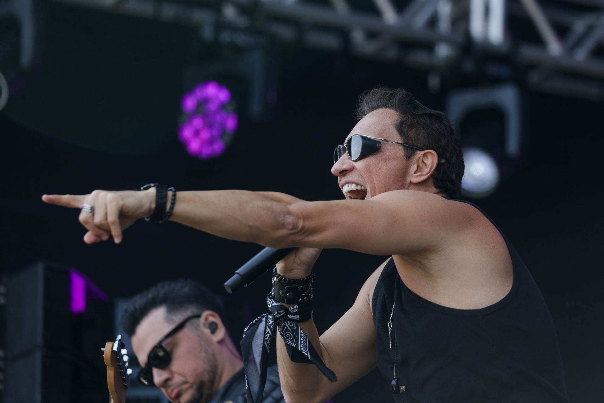 Um cantor de óculos escuros e regata preta aponta para a plateia enquanto se apresenta em um palco sob luzes de palco.