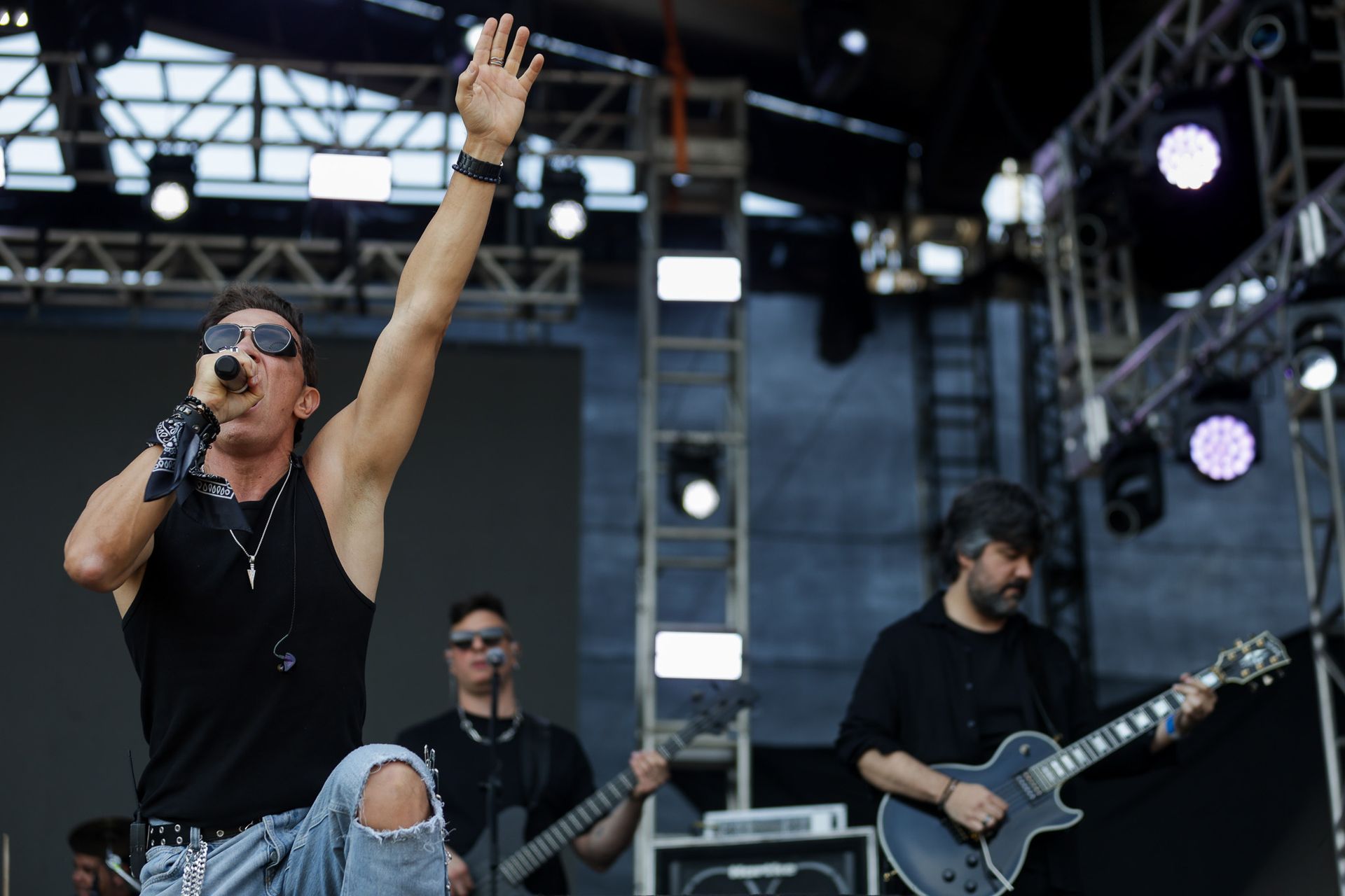 O vocalista gesticula enquanto se apresenta no palco com um guitarrista e um baixista atrás dele.