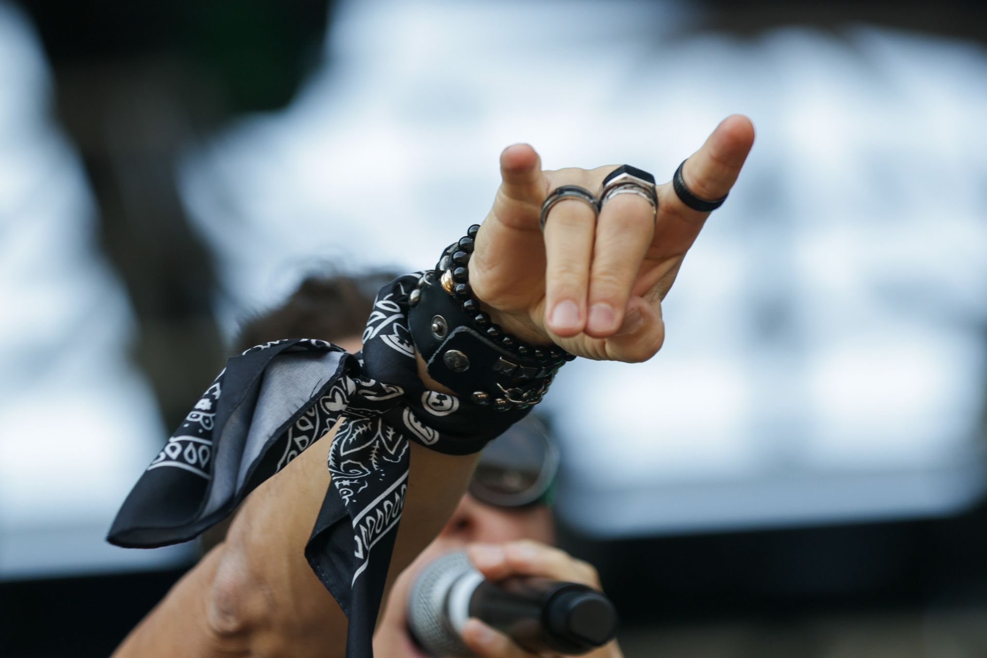 Um artista faz o gesto de mão característico do rock 'n' roll enquanto segura um microfone, usando uma bandana preta e anéis.