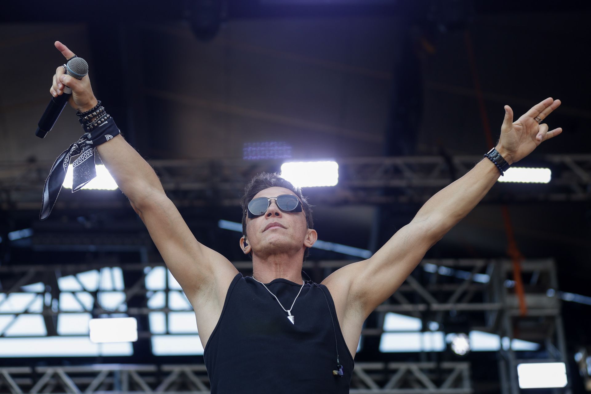Um artista vestindo uma regata preta e óculos escuros está no palco com os braços levantados, segurando um microfone.