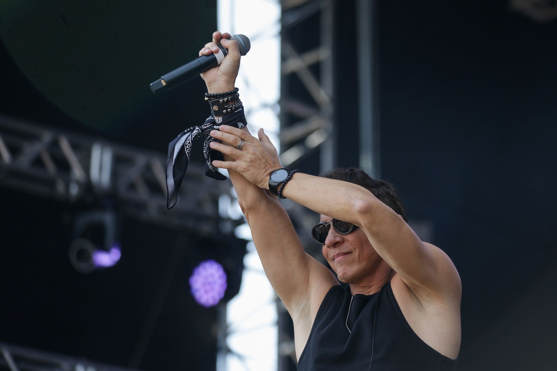 Um artista no palco segura um microfone bem acima da cabeça, usando óculos escuros e uma camiseta preta sem mangas.
