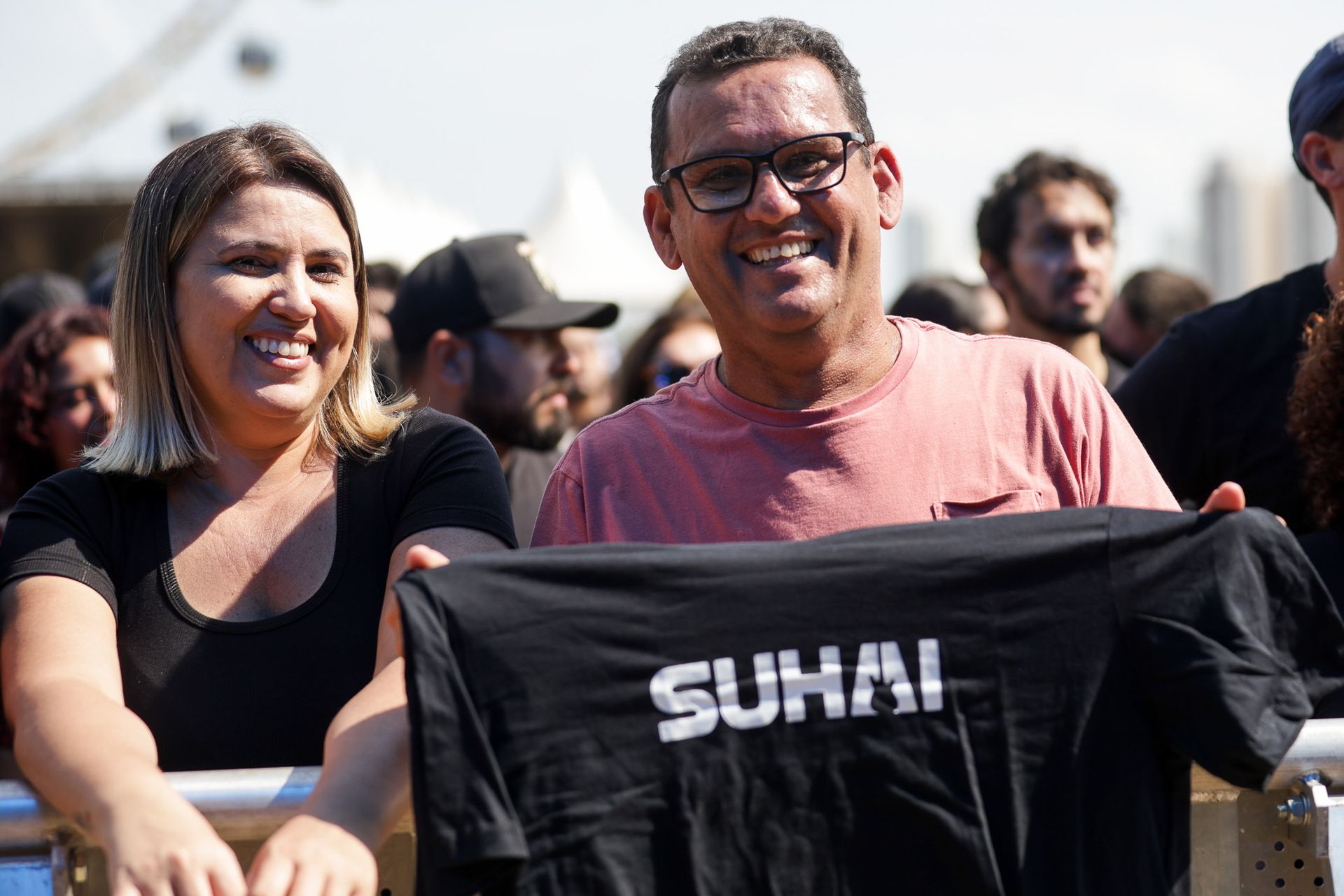 Um casal sorridente está em pé ao ar livre, segurando uma camiseta preta com a palavra 