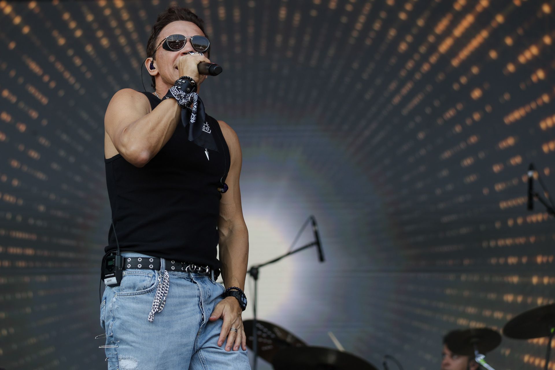 Um cantor vestindo uma regata preta e calça jeans se apresenta ao microfone em um palco com um fundo colorido e pontilhado.
