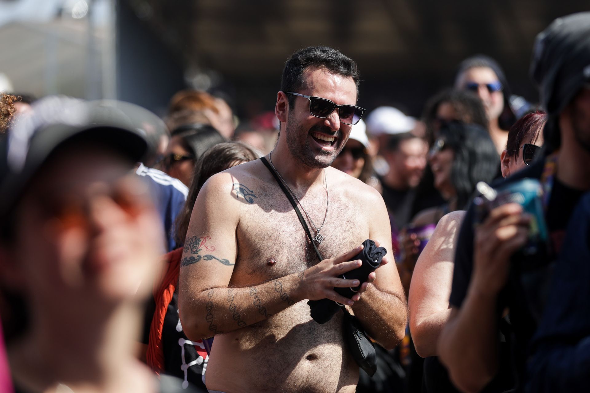 Uma pessoa sorridente, sem camisa, de óculos escuros e com tatuagens, segura uma câmera em um local externo movimentado.