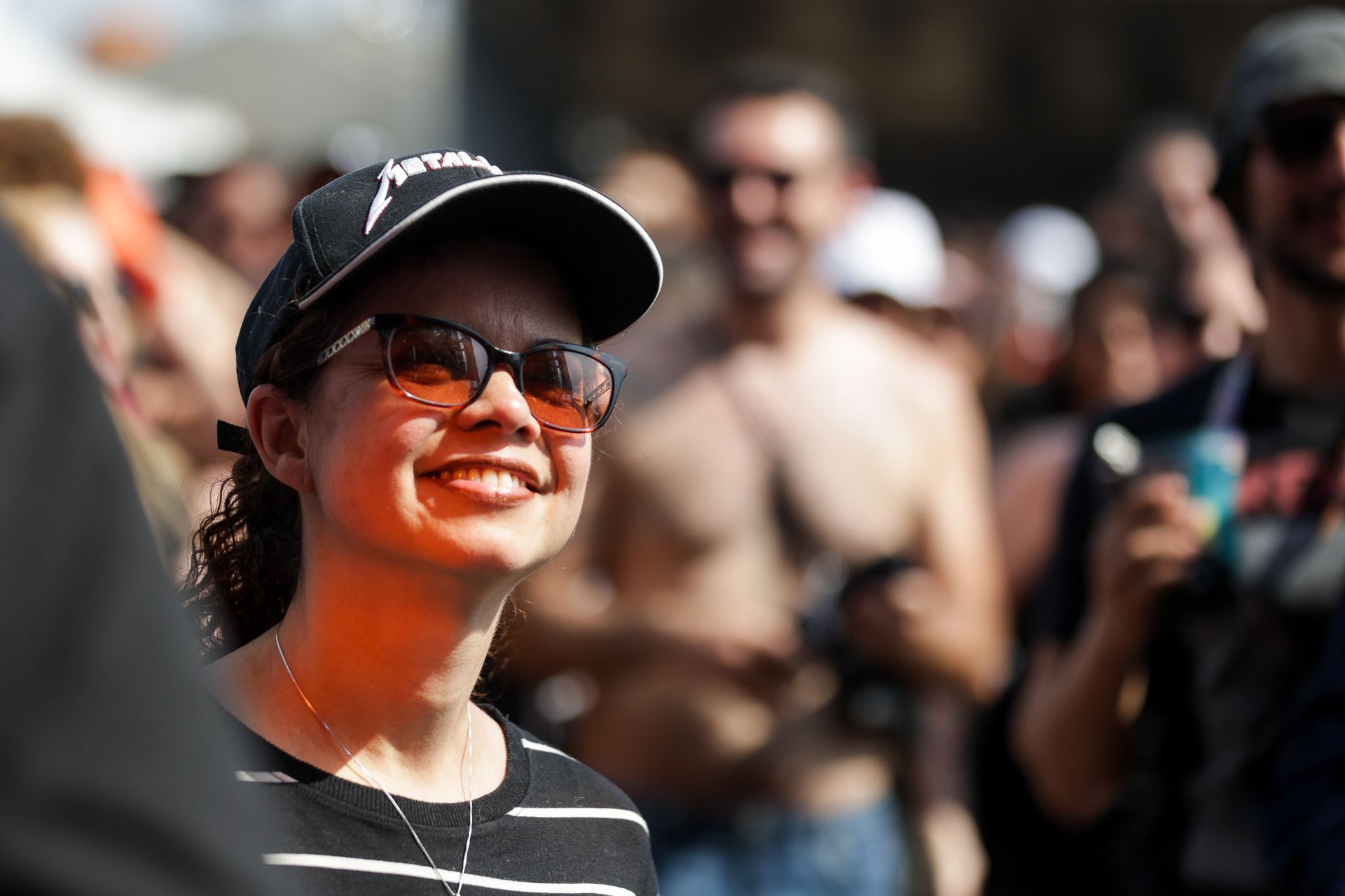 Pessoa sorridente com boné do Metallica e óculos escuros em um evento lotado ao ar livre, vista de lado.
