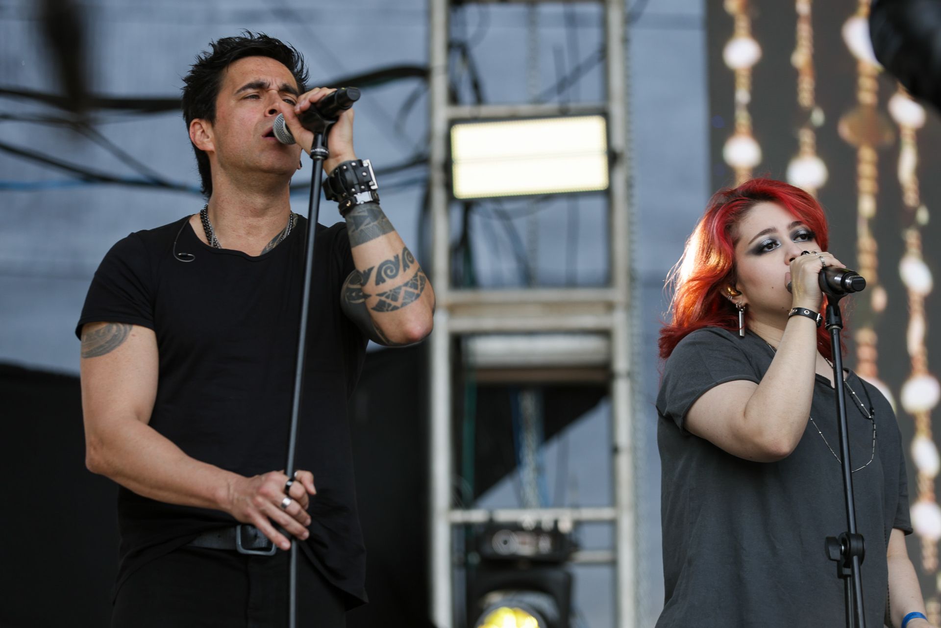 Duas pessoas cantando em microfones em um palco de shows com uma estrutura de iluminação metálica ao fundo.