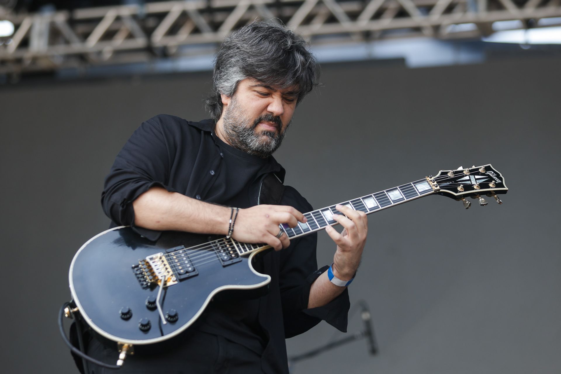 Um músico de cabelos escuros e barba toca uma guitarra no palco, concentrando-se intensamente.
