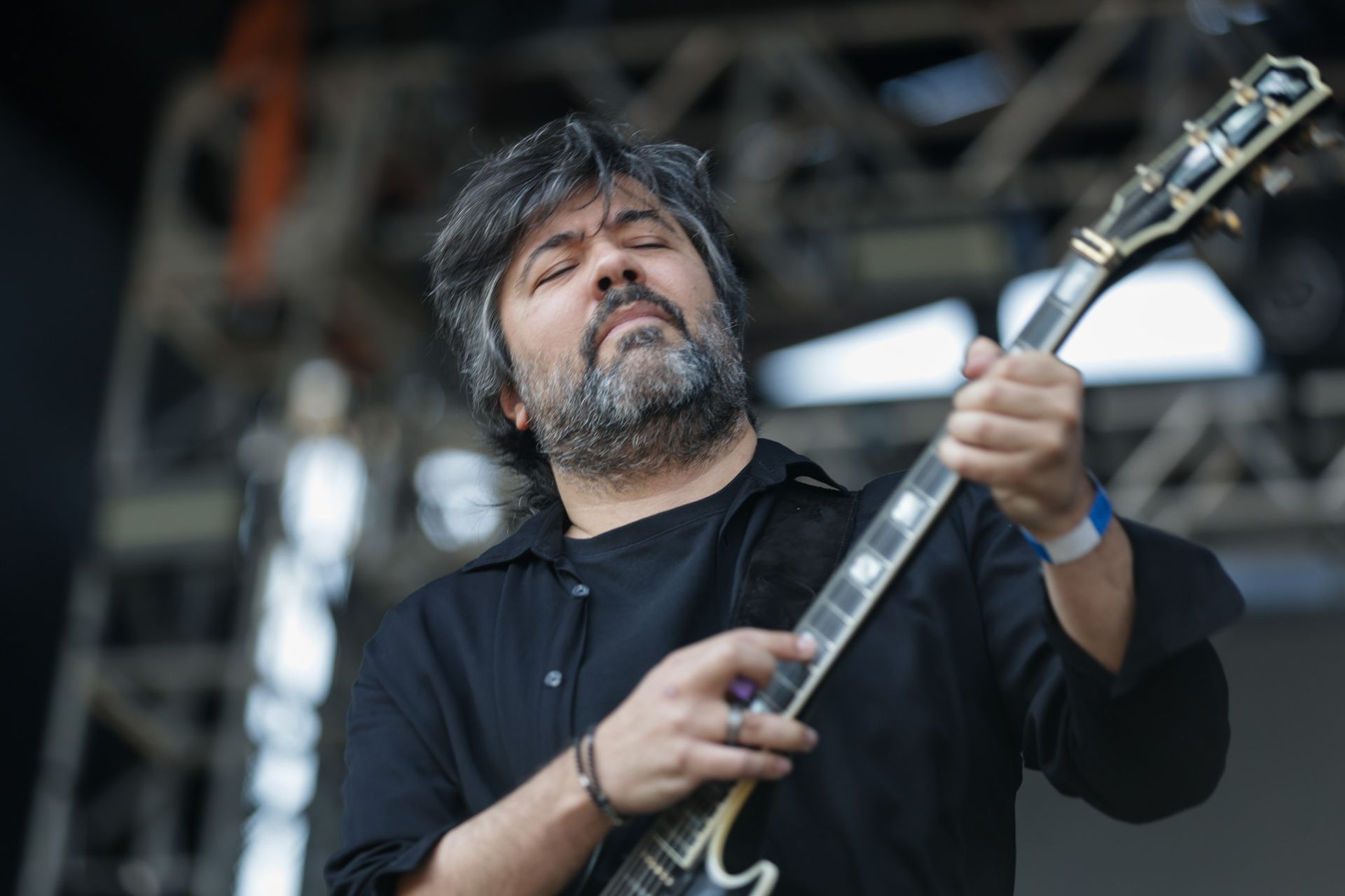 Uma pessoa com barba, vestindo uma camisa preta, tocando guitarra elétrica ao ar livre com os olhos fechados, concentrada.