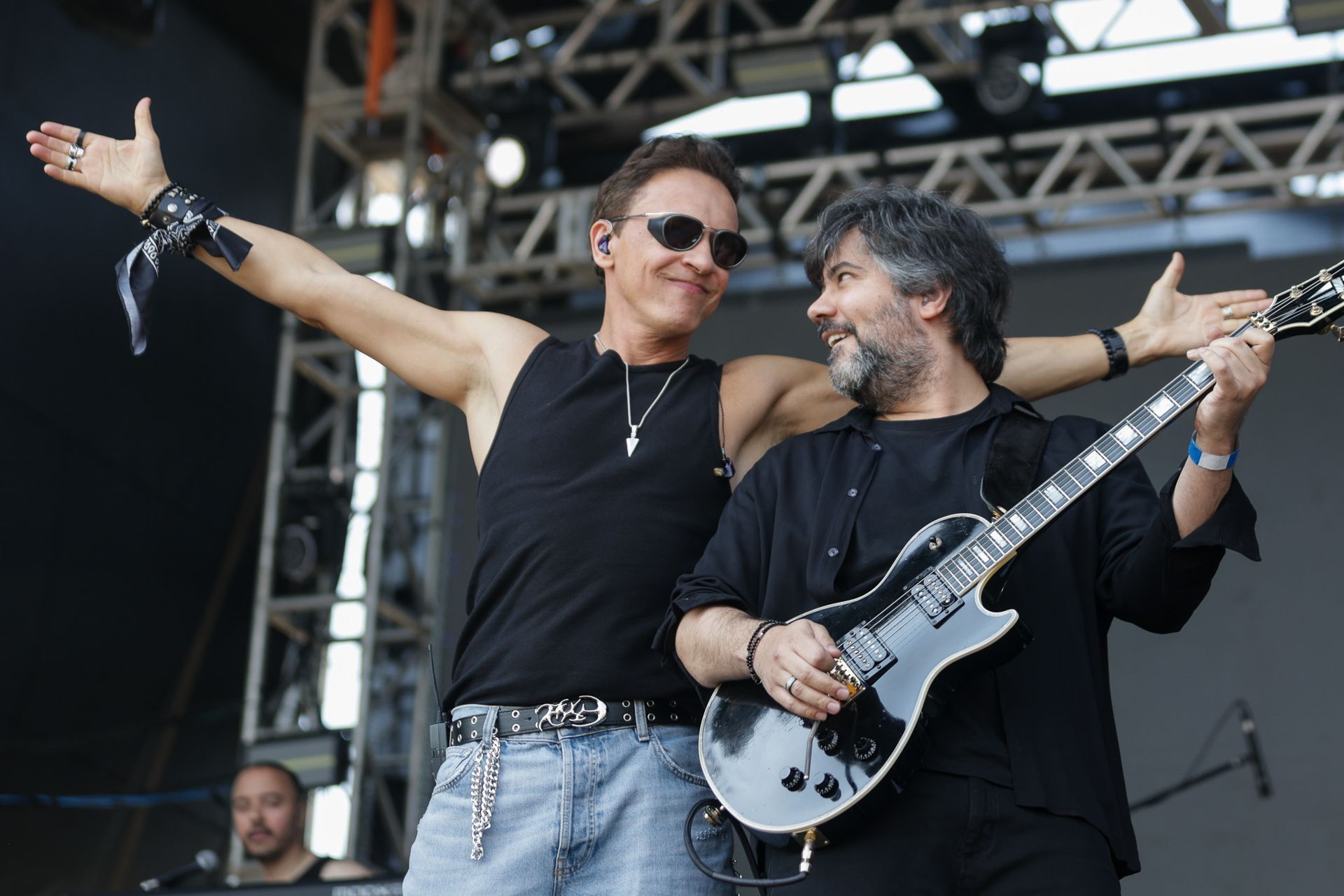 Dois músicos se apresentando em um palco; um está de pé com os braços estendidos enquanto o outro sorri e toca uma guitarra.