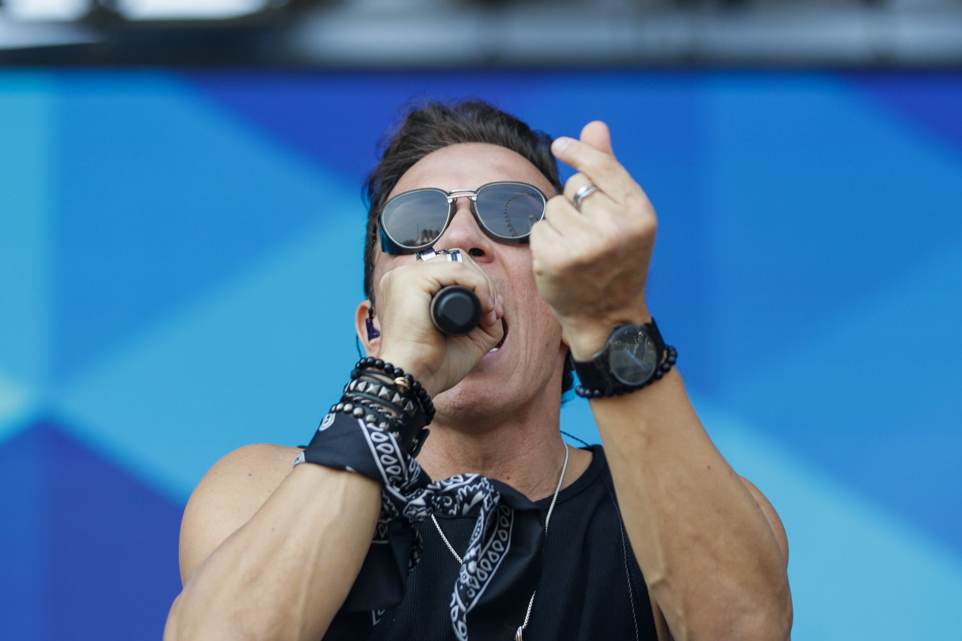 Um cantor se apresentando no palco, usando óculos escuros e uma bandana enquanto gesticula com uma das mãos.