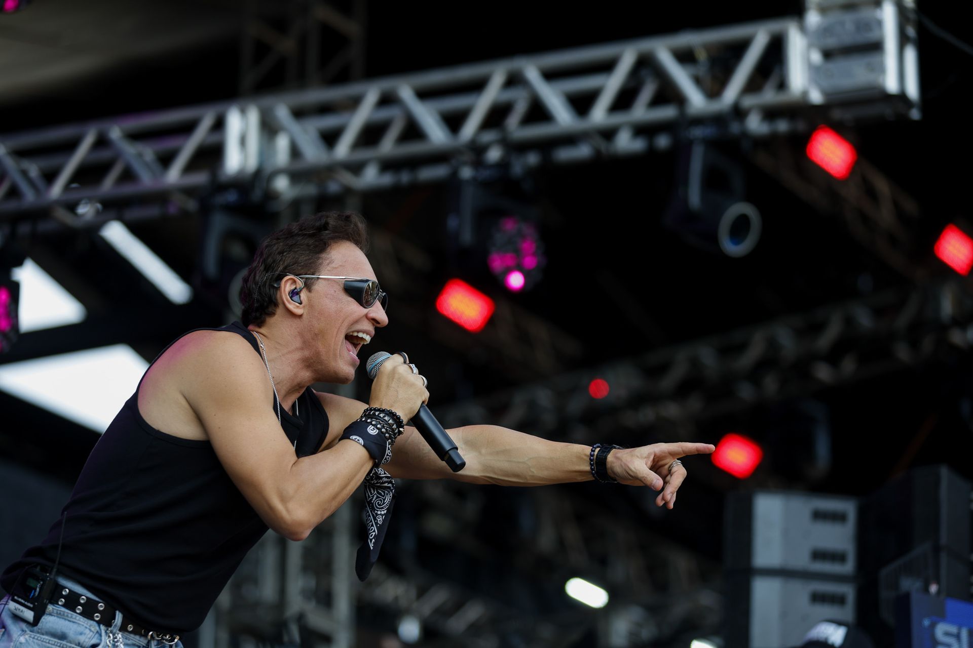 Um cantor de regata preta e óculos escuros se apresenta no palco, apontando para a frente enquanto segura um microfone.