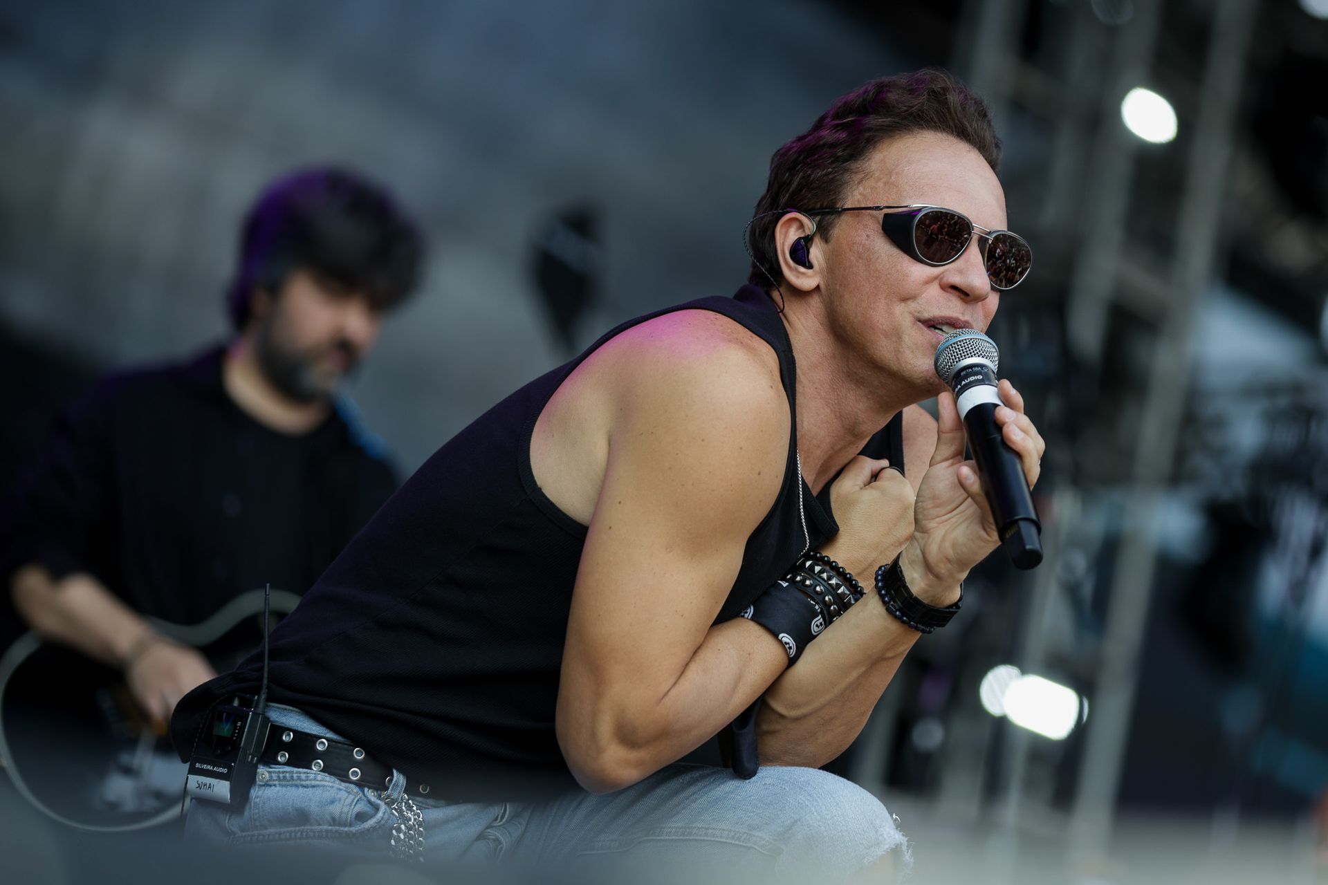 Um artista de regata preta e óculos escuros se apresentando no palco com um microfone, com um guitarrista desfocado ao fundo.