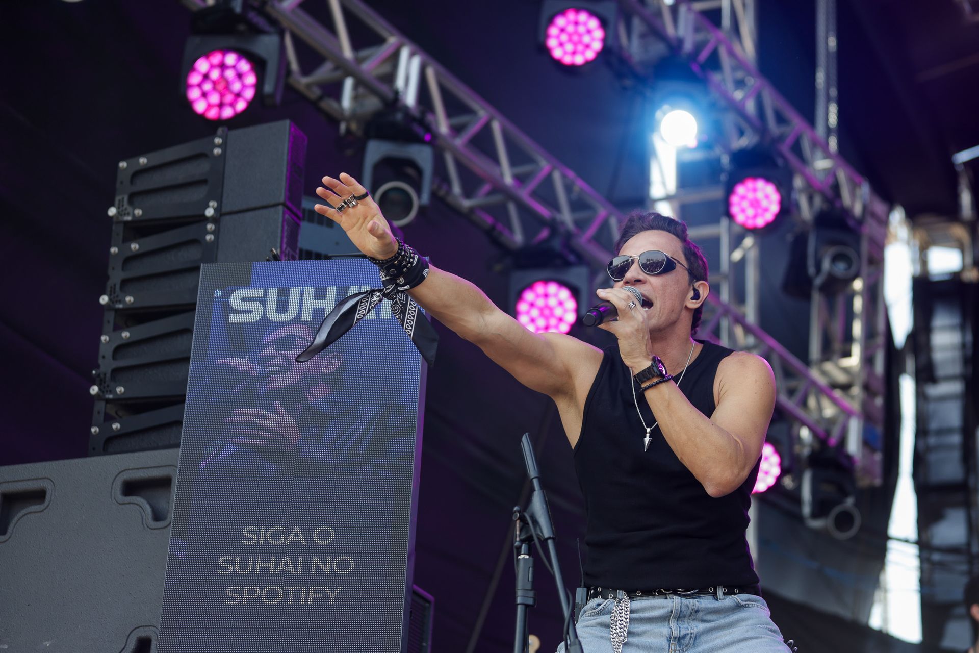 Um músico vestindo uma regata preta e óculos escuros canta enquanto estende a mão em direção à plateia no palco.